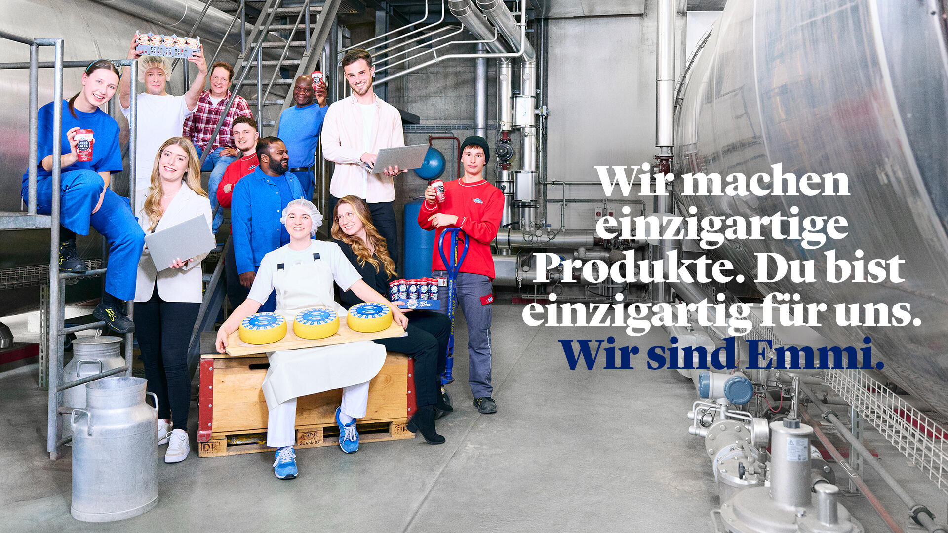Eine bunt gemischte Gruppe lächelnder Arbeiter posiert in einer Molkerei mit Laptops, Käselaiben und Tabletts mit Produkten in der Hand. Der deutsche Text lautet: Wir sind Emmi. Wir machen einzigartige Produkte. Du bist einzigartig für uns.