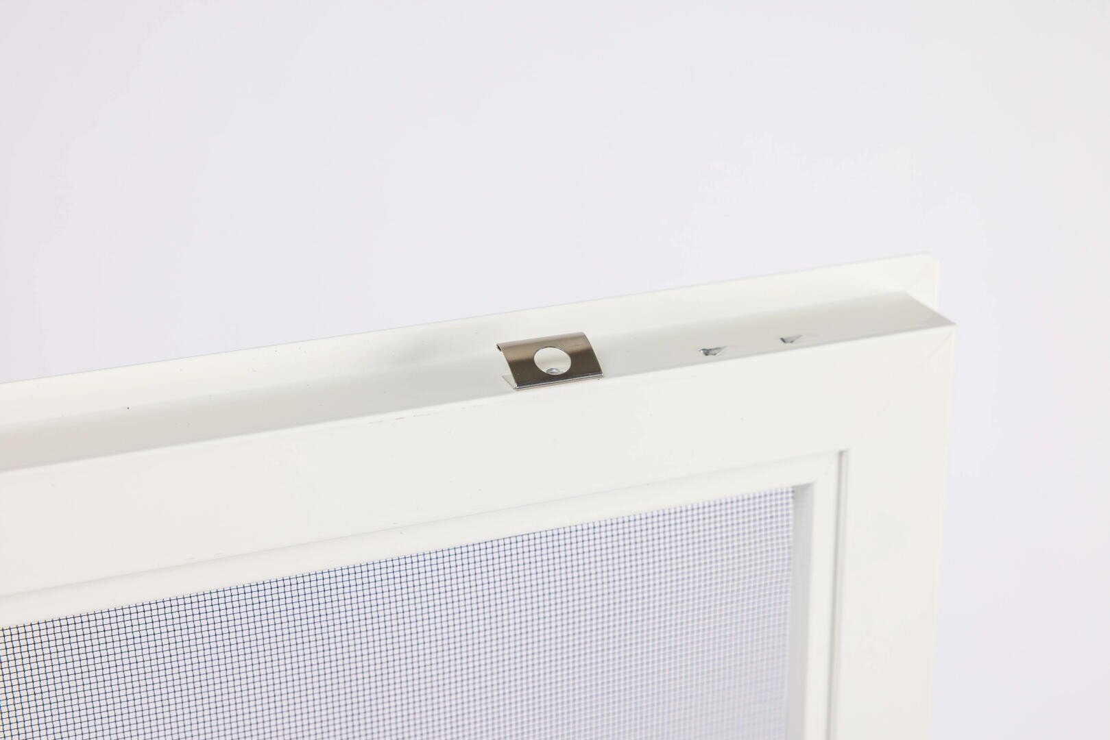 Nahaufnahme der oberen Kante eines weißen Fenstergitters mit einer kleinen Metallklammer, die am Rahmen befestigt ist, vor einem schlichten weißen Hintergrund.
