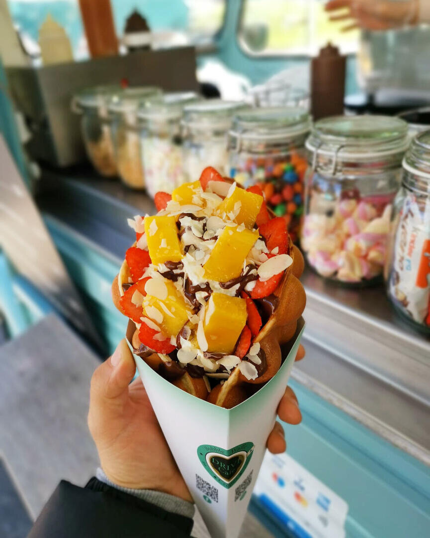 Eine Hand hält eine mit Erdbeerscheiben, Mangostückchen, Schokoladensauce und Mandelblättchen gefüllte Waffeltüte, während im Hintergrund Gläser mit bunten Toppings zu sehen sind.