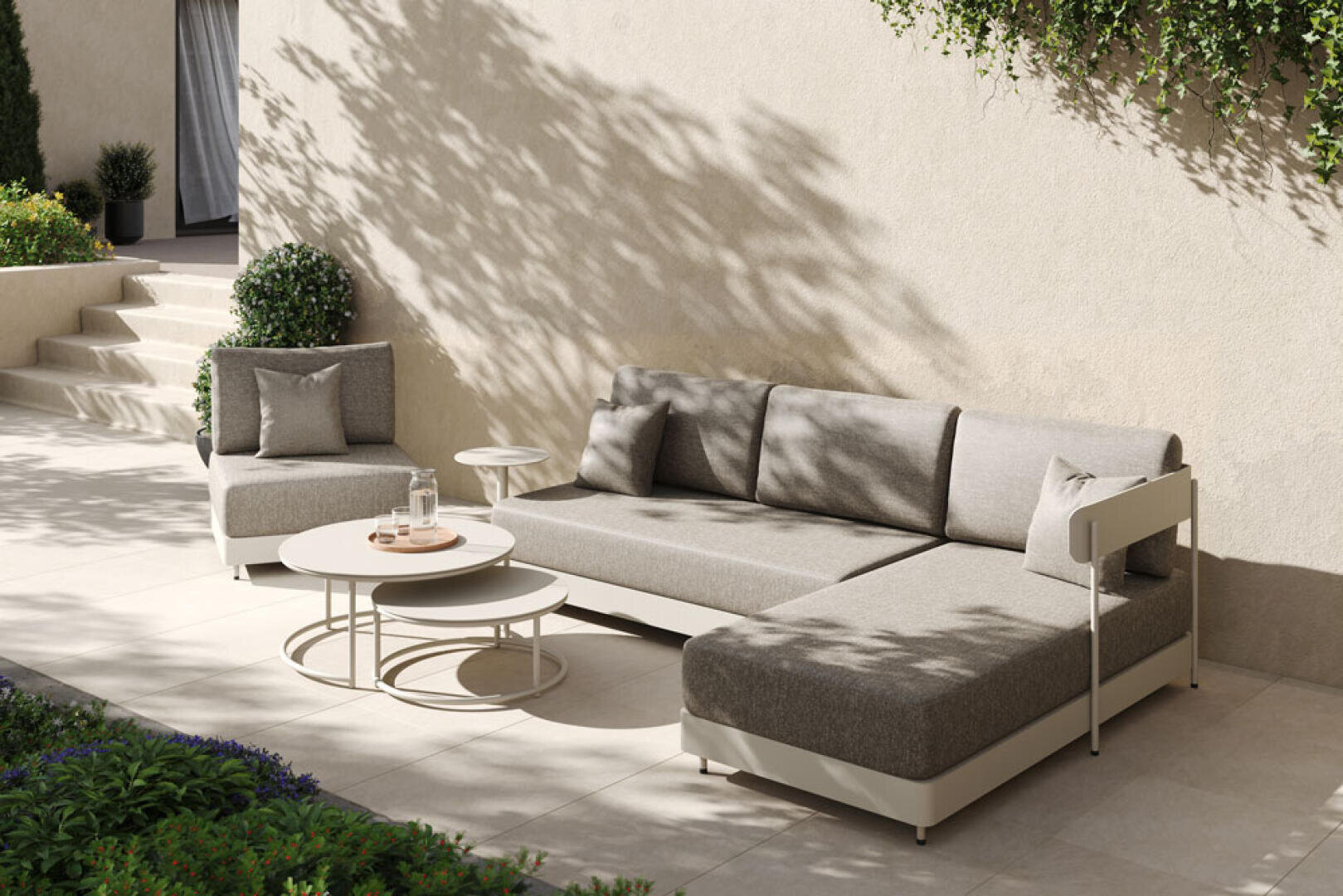 Eine moderne Außenterrasse mit einem beigen L-förmigen Sofa, einem passenden Sessel, runden Couchtischen mit Gläsern und üppigen Grünpflanzen vor einer hellen Stuckwand im gedämpften Sonnenlicht.