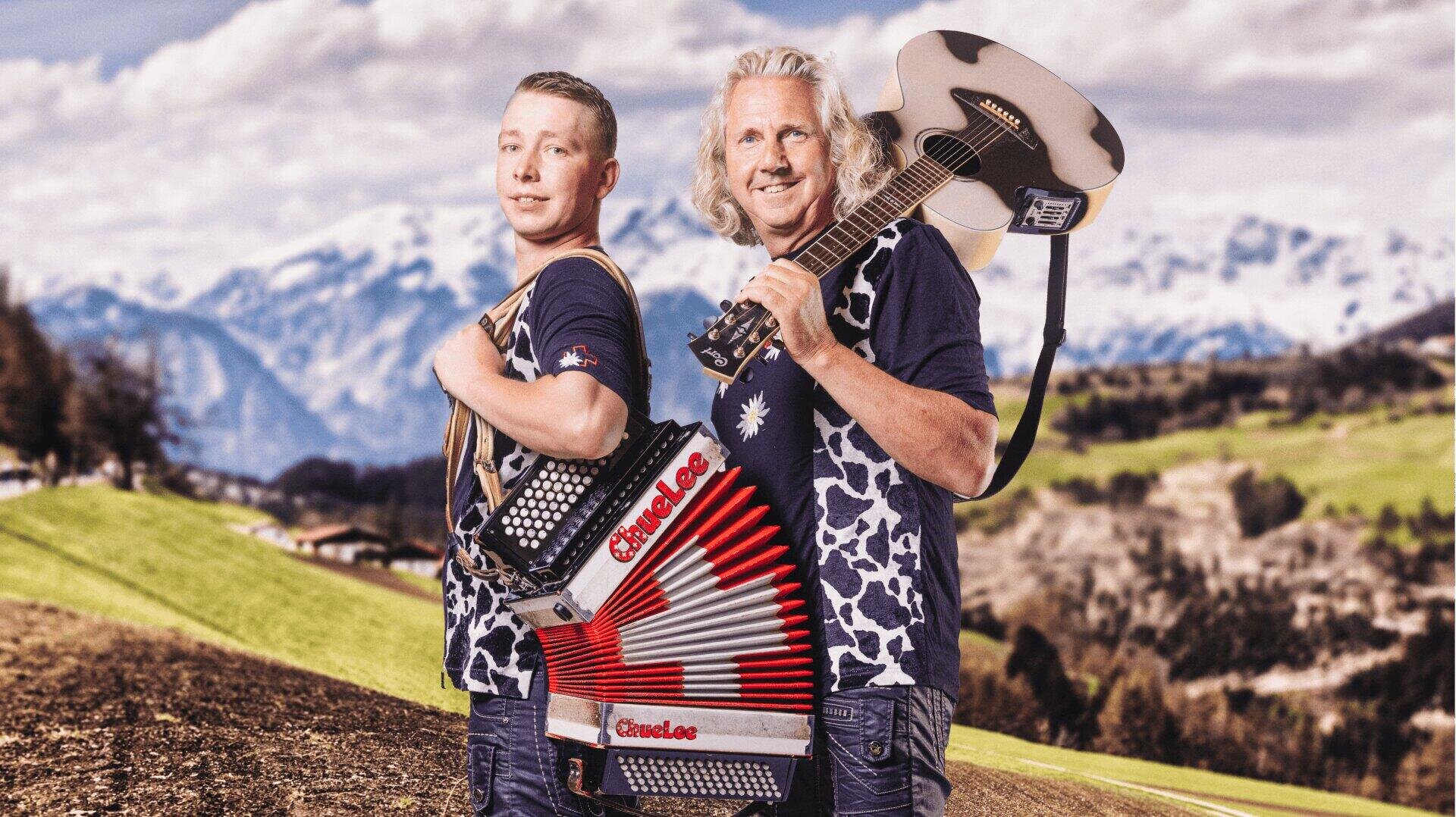 Zwei Männer stehen im Freien in einer Berglandschaft, der eine hält ein Akkordeon, der andere eine Gitarre. Beide lächeln und tragen blaue Hemden mit weißen Mustern. Hinter ihnen sind schneebedeckte Berge und grüne Felder zu sehen.