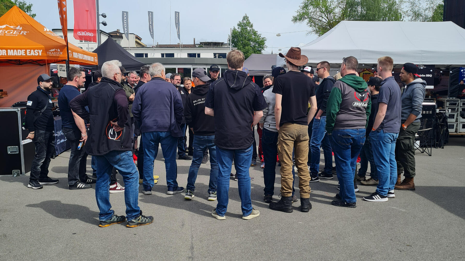 Eine Gruppe von Menschen steht in einem Kreis im Freien auf dem Bürgersteig, umgeben von Zelten und Veranstaltungsständen. Die meisten tragen Freizeitkleidung und Jacken, und das Wetter scheint mild und sonnig zu sein.