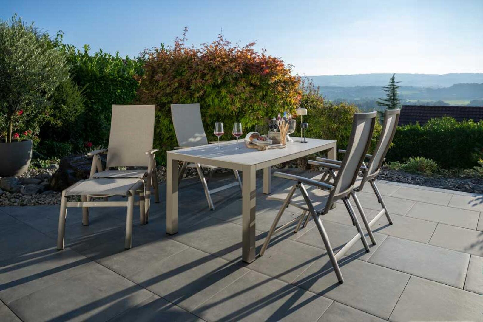 Moderne Außenterrasse mit einem weißen Esstisch für vier Personen, umgeben von sechs grauen Stühlen. Auf dem Tisch stehen Weingläser, Snacks und Kerzen, während im Hintergrund üppiges Grün und Hügel zu sehen sind.