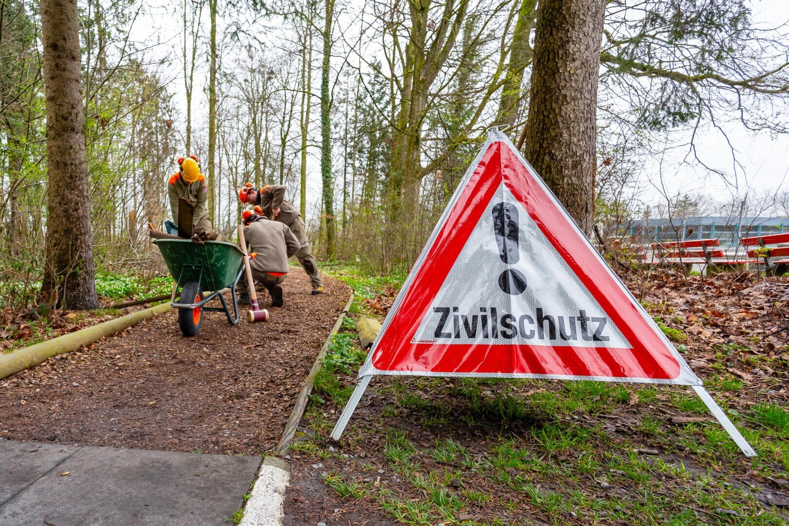 Ein dreieckiges Warnschild mit der Aufschrift "Zivilschutz" steht an einem Weg in einem Waldgebiet, während im Hintergrund Personen in Schutzkleidung mit einer Schubkarre arbeiten.