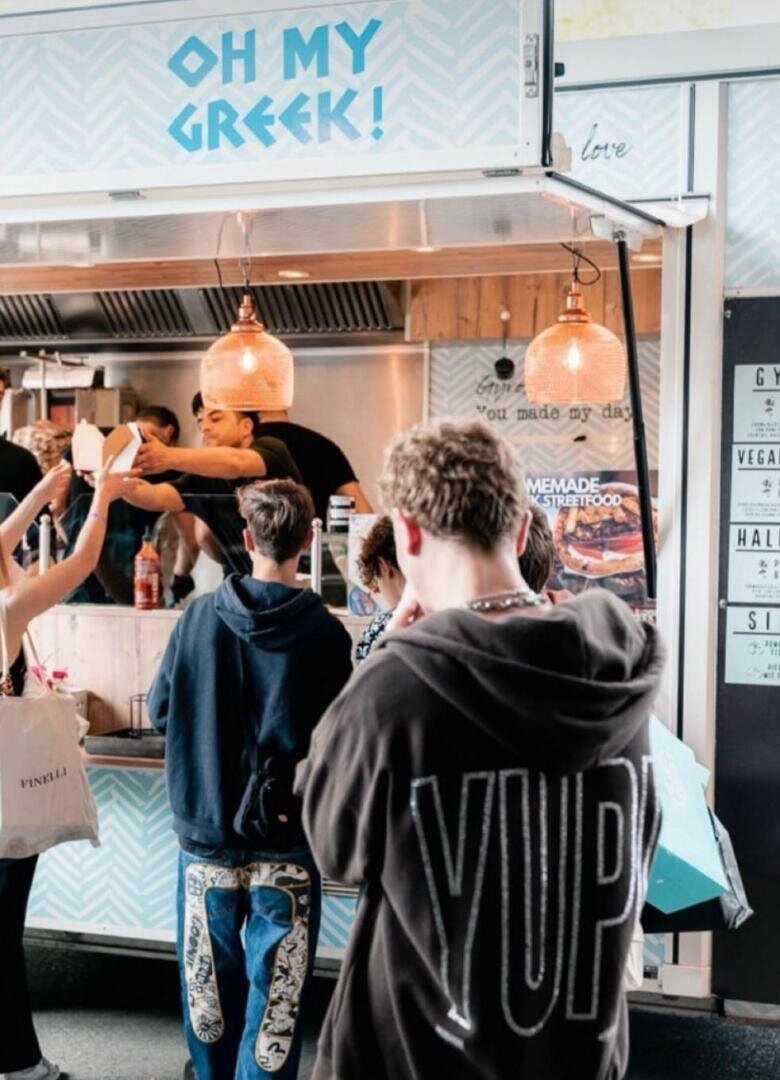 Die Leute stehen in der Schlange vor einem Food Truck mit dem Namen OH MY GREEK! während ein Mitarbeiter im Inneren Essen ausgibt. Der Wagen hat Hängelampen und eine Speisekarte mit griechischen Gerichten. Ein Kunde trägt einen Kapuzenpulli mit der Aufschrift YUP auf dem Rücken.