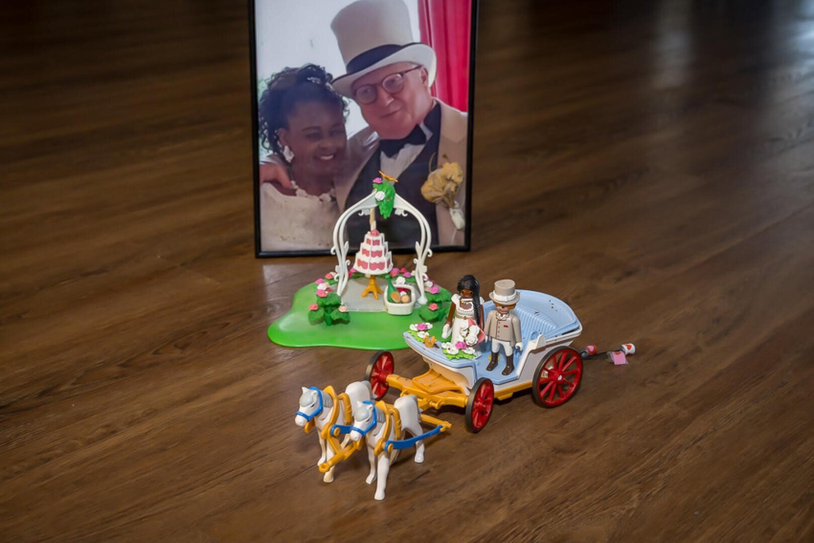 Ein gerahmtes Foto eines lächelnden Paares in Hochzeitskleidung steht auf einem Holzboden hinter Spielzeug-Hochzeitsfiguren, einer dekorierten Torte und einer Pferdekutsche.