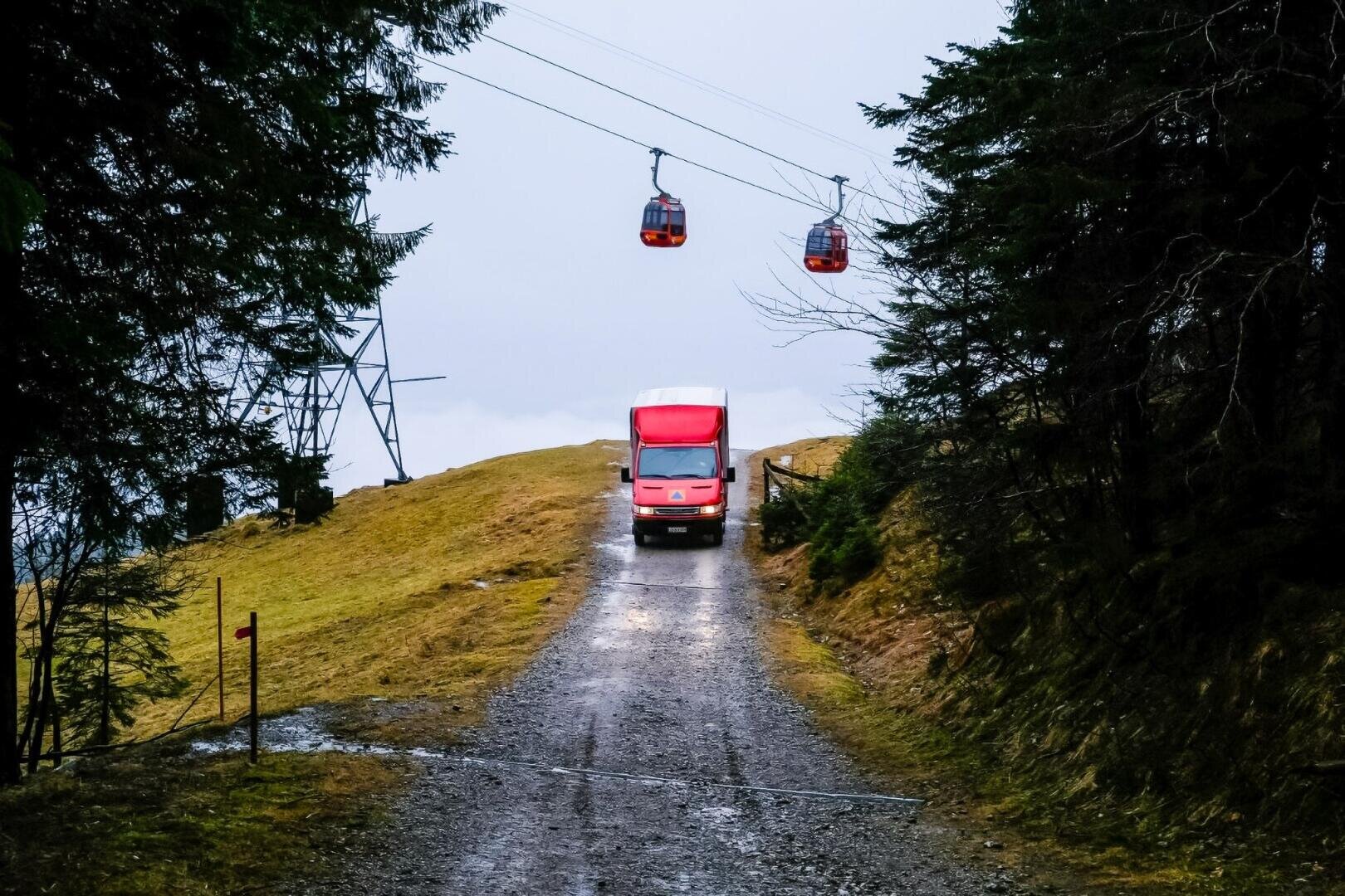 Ein roter Lieferwagen fährt einen schmalen, von Bäumen gesäumten Schotterweg hinauf, während drei rote Seilbahnen vor einem wolkenverhangenen Himmel vorbeifahren. Der Boden ist nass, und die Szene scheint in einer hügeligen oder bergigen Gegend zu spielen.