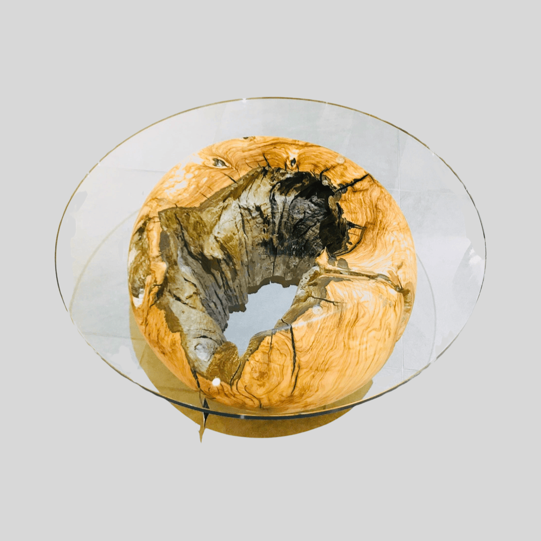 Ein runder Couchtisch aus Glas mit einem großen, ausgehöhlten Holzsockel mit natürlicher, rauer Textur und sichtbarer Holzmaserung vor einem schlichten grauen Hintergrund.