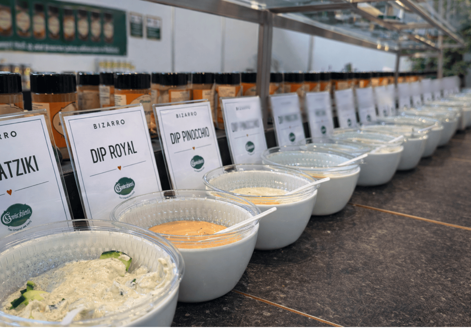 Eine Reihe weißer Schalen mit verschiedenen Dips, die jeweils mit Aufschriften wie Tzatziki, Dip Royal und Dip Pinocchio versehen sind, stehen auf einer Arbeitsplatte mit Gewürzdosen im Hintergrund.