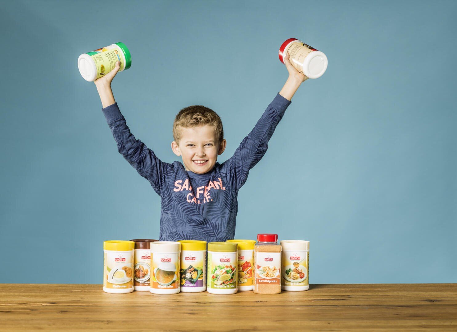 Ein lächelnder Junge mit erhobenen Armen hält zwei Gewürzdosen und steht hinter einer Reihe verschiedener Gewürzdosen auf einem Holztisch mit einfarbig blauem Hintergrund.