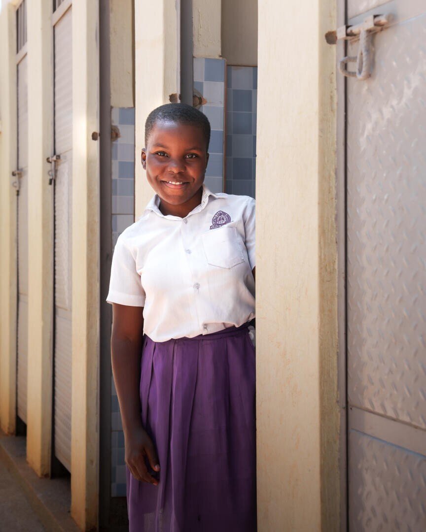 Ein lächelndes Mädchen in einem weißen Hemd und einem lila Rock steht in einem Korridor neben Metall- und Kacheltüren, möglicherweise in einer Schule.