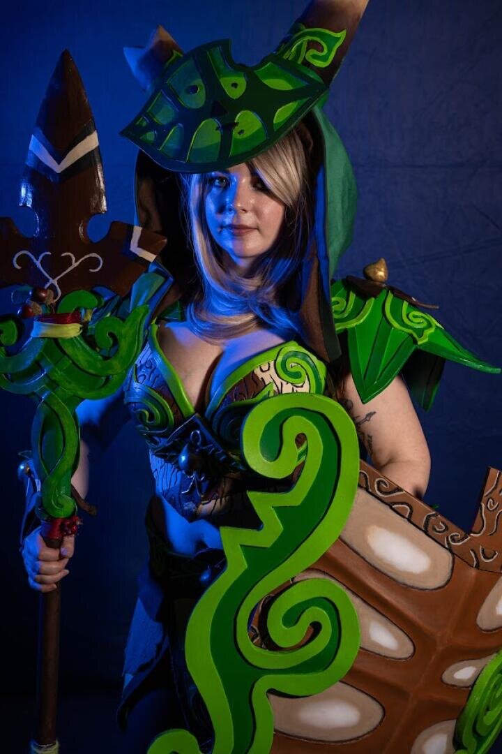 Eine Person in aufwändiger grüner und brauner Fantasierüstung mit Blattmustern hält einen großen Stab und ein Schild und posiert vor einem dunkelblauen Hintergrund. Das Kostüm weist aufwändige Details auf, darunter einen Helm und Schulterstücke.