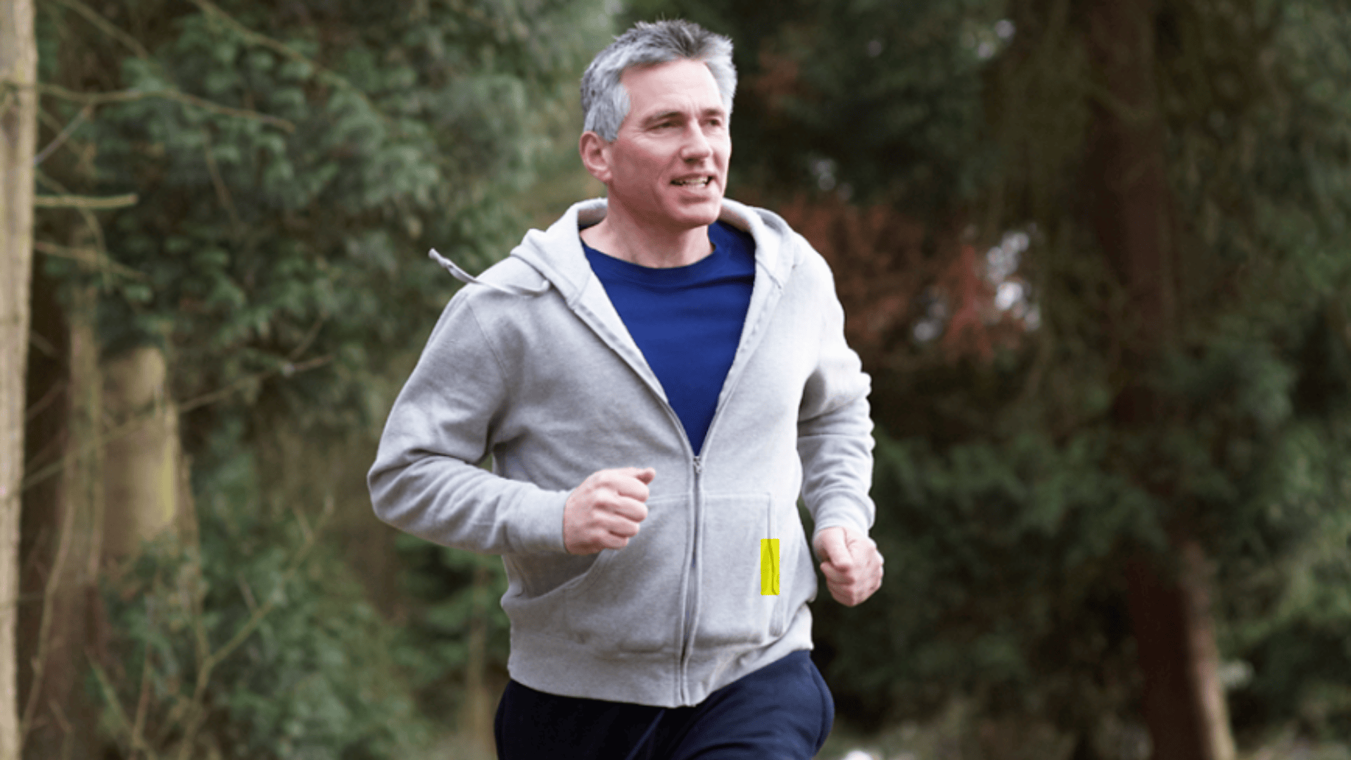 Ein Mann mittleren Alters joggt im Freien durch ein bewaldetes Gebiet, er trägt einen grauen Kapuzenpullover mit Reißverschluss und eine dunkle Hose. Er wirkt konzentriert und zufrieden, mit Bäumen und Grünzeug im Hintergrund.