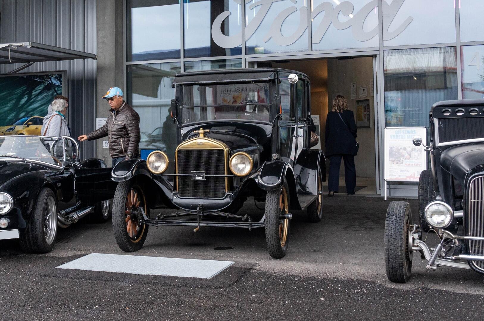 Ein alter schwarzer Ford ist vor einem Gebäude mit einem großen Ford-Schild geparkt. Drei Personen stehen in der Nähe, und zwei weitere Oldtimer sind teilweise auf beiden Seiten des Fords zu sehen.