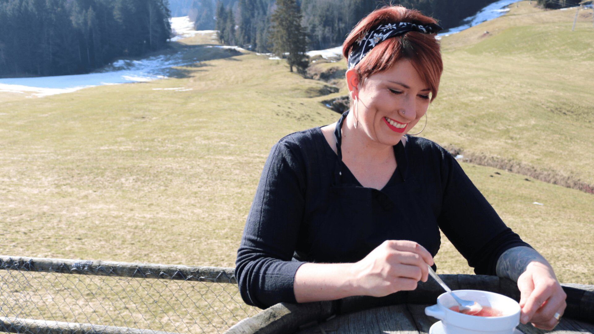 Eine Frau mit kurzen roten Haaren und einem gemusterten Stirnband sitzt im Freien an einem Holzgeländer und rührt lächelnd in einer Schüssel mit Essen. In der grasbewachsenen Landschaft hinter ihr sind Schneeflecken und Bäume zu sehen.