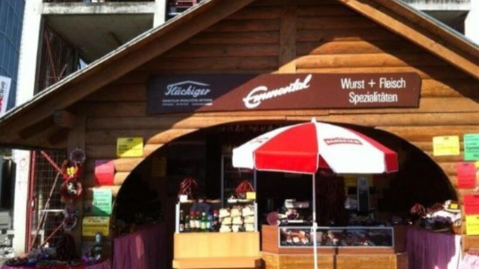 Ein hölzerner Marktstand verkauft Wurst- und Fleischspezialitäten, mit einem rot-weißen Schirm davor und bunten Schildern an den Wänden. Der Stand befindet sich in einem sonnigen Außenbereich.