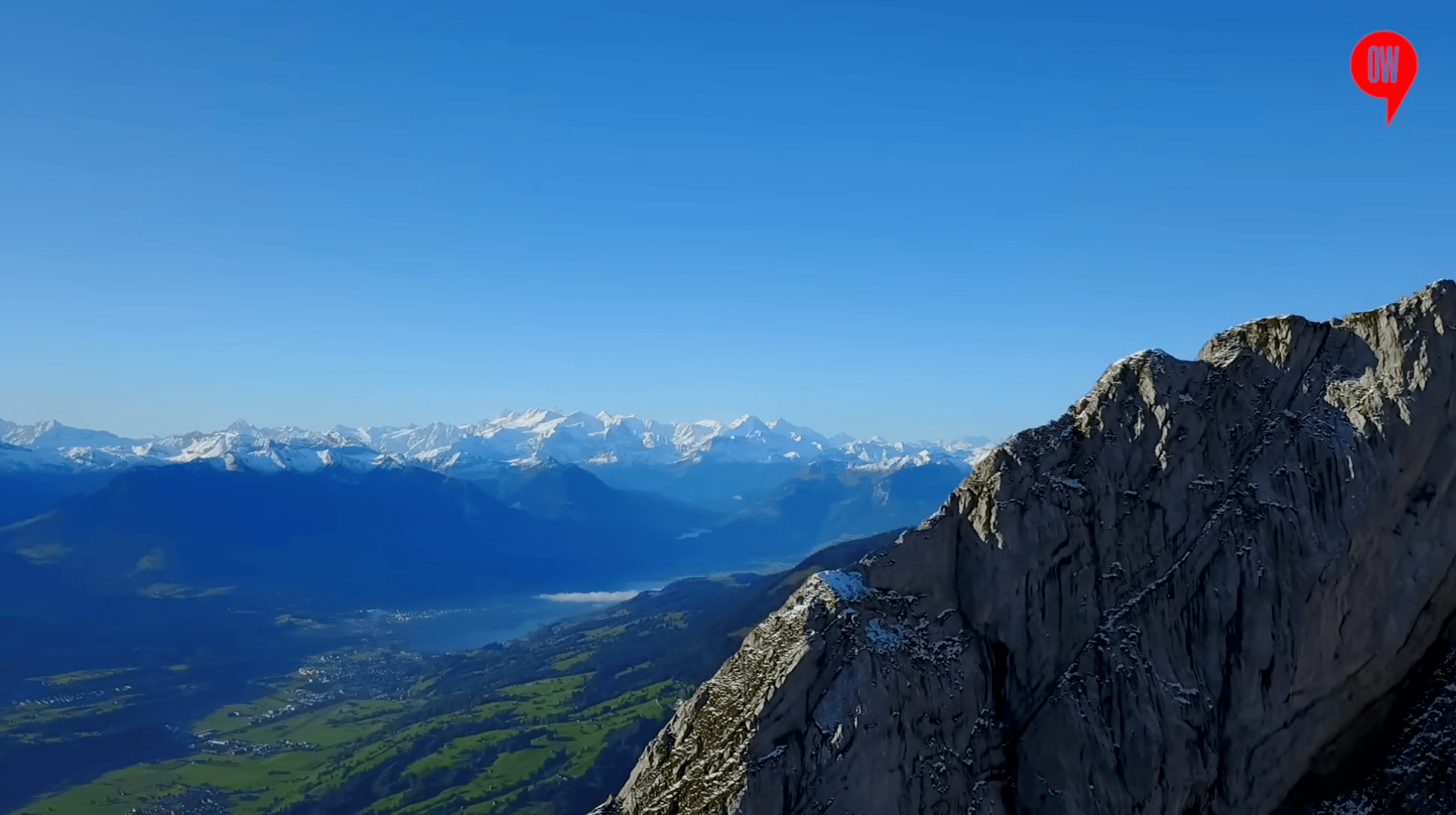 Ein Panoramablick auf einen zerklüfteten Berggipfel im Vordergrund mit schneebedeckten Bergen und einem klaren blauen Himmel in der Ferne. Darunter liegt ein Tal mit Grün und einem See.