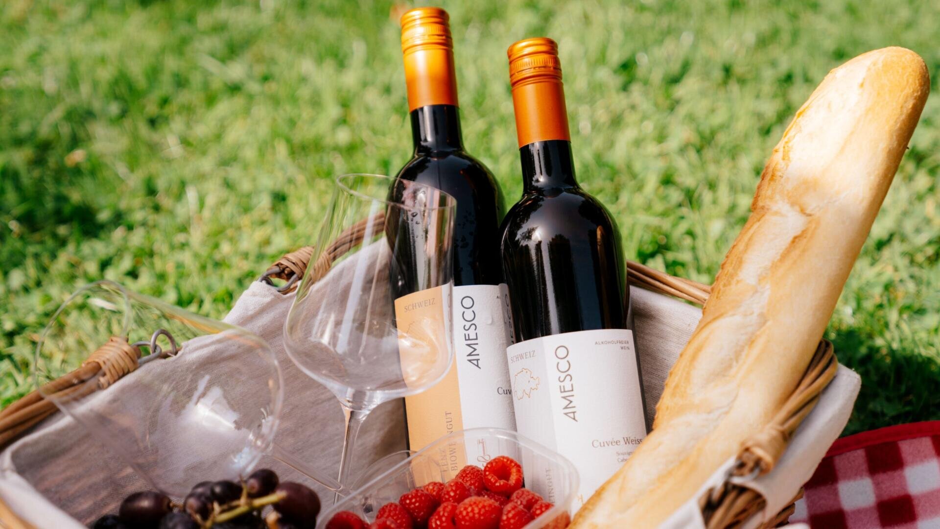 Ein Picknickkorb im Gras enthält zwei Flaschen Rotwein, zwei leere Weingläser, ein Baguette, rote Weintrauben und Erdbeeren. Die Szene erinnert an ein entspanntes Essen im Freien.
