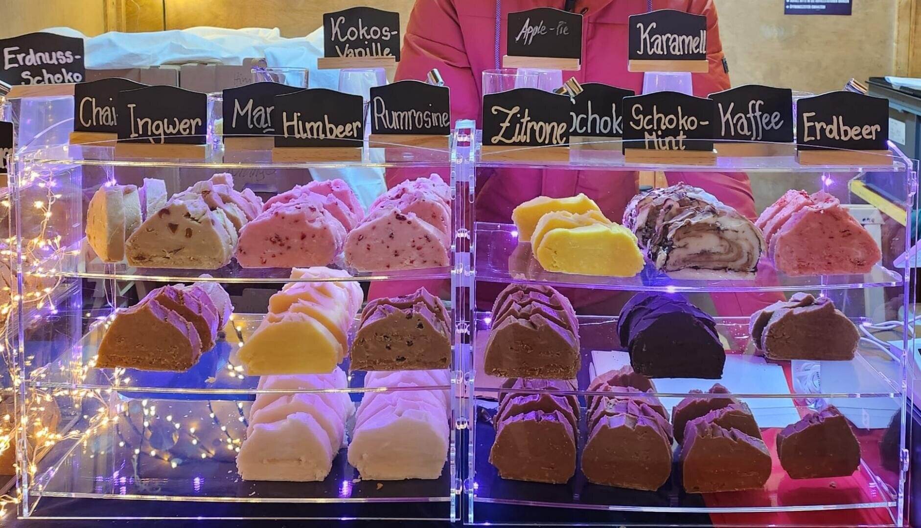 In einer Vitrine stehen bunte, dreieckige Gebäckstücke mit Geschmacksrichtungen wie Chai, Marzipan, Rum-Rosinen, Karamell, Zitrone, Kaffee und Erdbeere, mit Lichterketten und einer Person, die hinter der Theke steht.