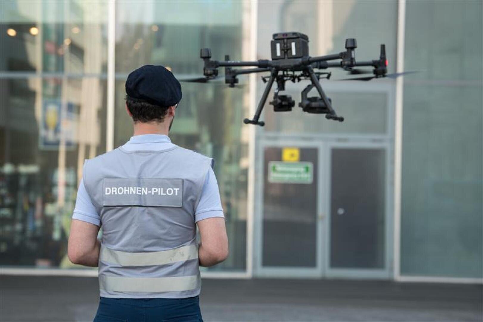 Eine Person, die eine Weste mit der Aufschrift DROHNEN-PILOT trägt, steuert eine große Drohne außerhalb eines modernen Gebäudes mit Glastüren und Fenstern.