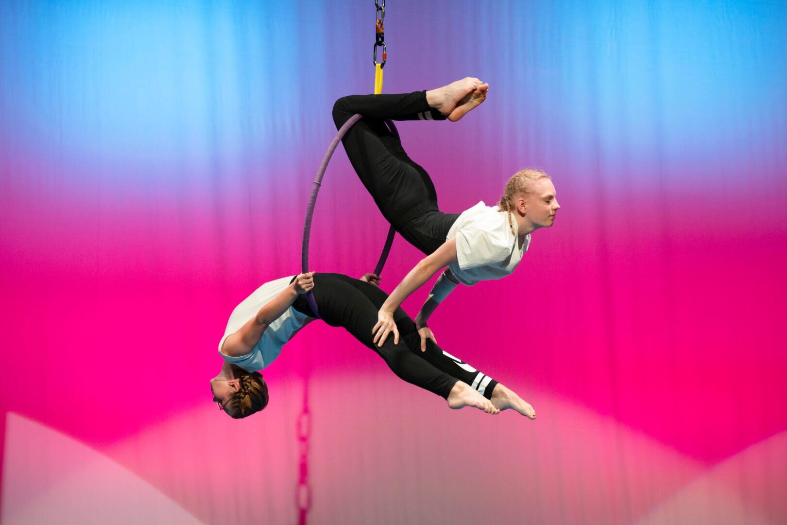 Zwei Luftakrobaten balancieren auf einem aufgehängten Reifen, jeder hält eine Pose mit gewölbtem Körper und ausgestreckten Beinen vor einem Hintergrund mit leuchtenden rosa und blauen Farbverläufen.