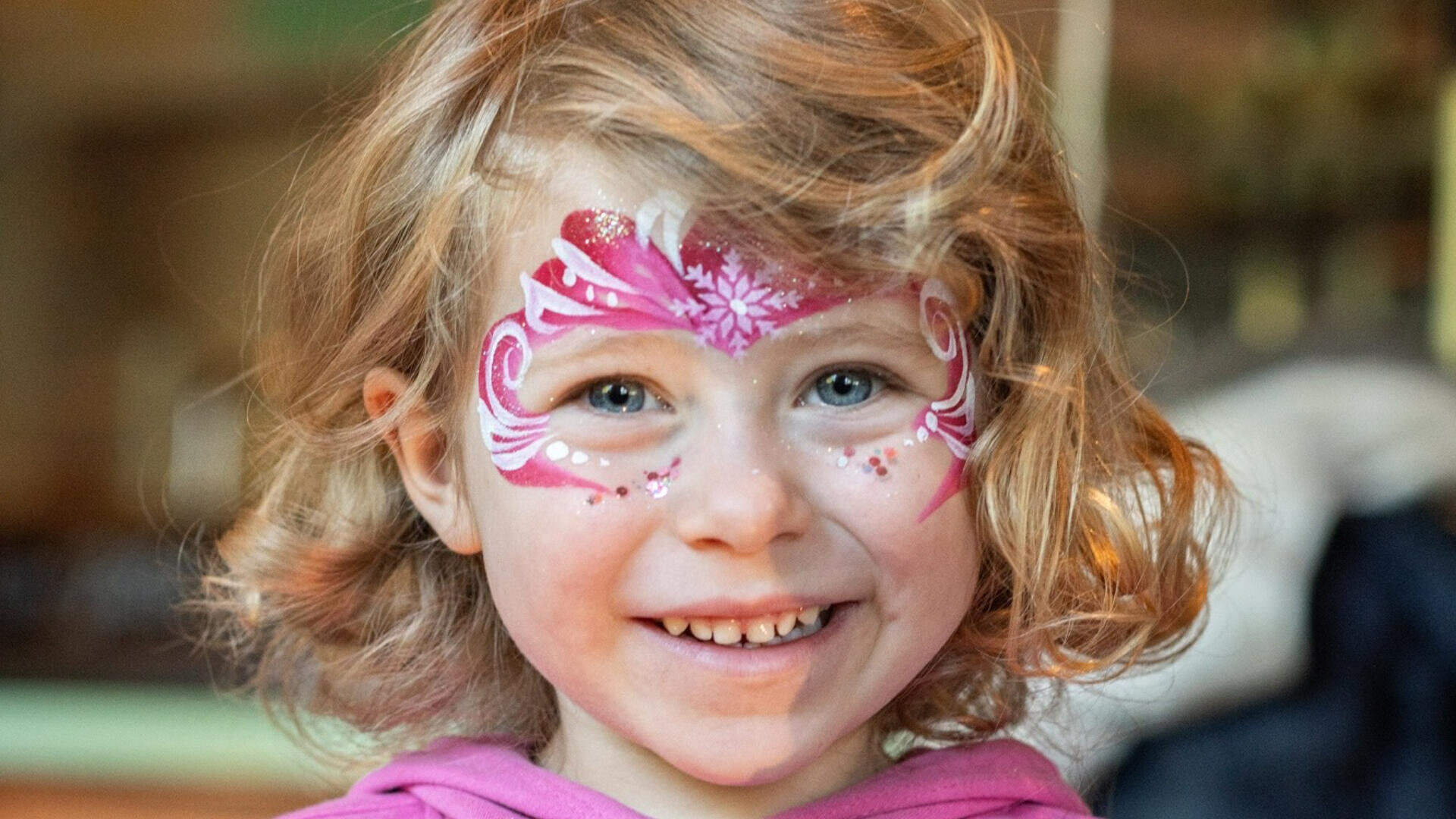 Ein lächelndes kleines Kind mit hellbraunem, lockigem Haar trägt eine rosa und weiße Gesichtsbemalung mit Glitzer, die an einen Schmetterling oder eine Maske erinnert. Das Kind ist mit einem rosa Oberteil bekleidet und befindet sich in einem Haus.