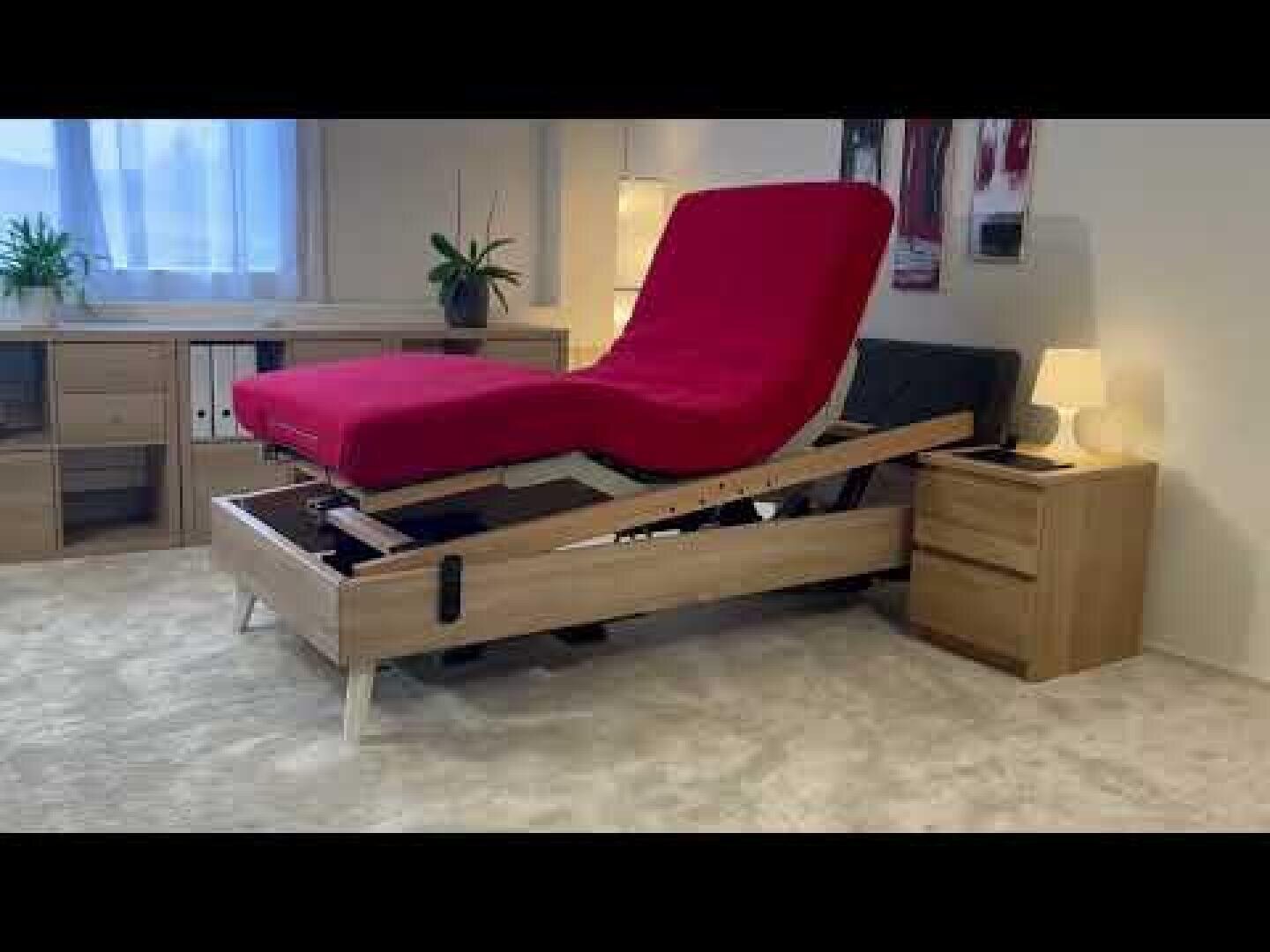 Ein verstellbares Holzbett mit einer leuchtend roten Matratze ist am Kopf und an den Füßen hochgezogen. Das Bett steht in einem modernen, gut beleuchteten Schlafzimmer mit Nachttischen, Lampen und Pflanzen im Hintergrund.