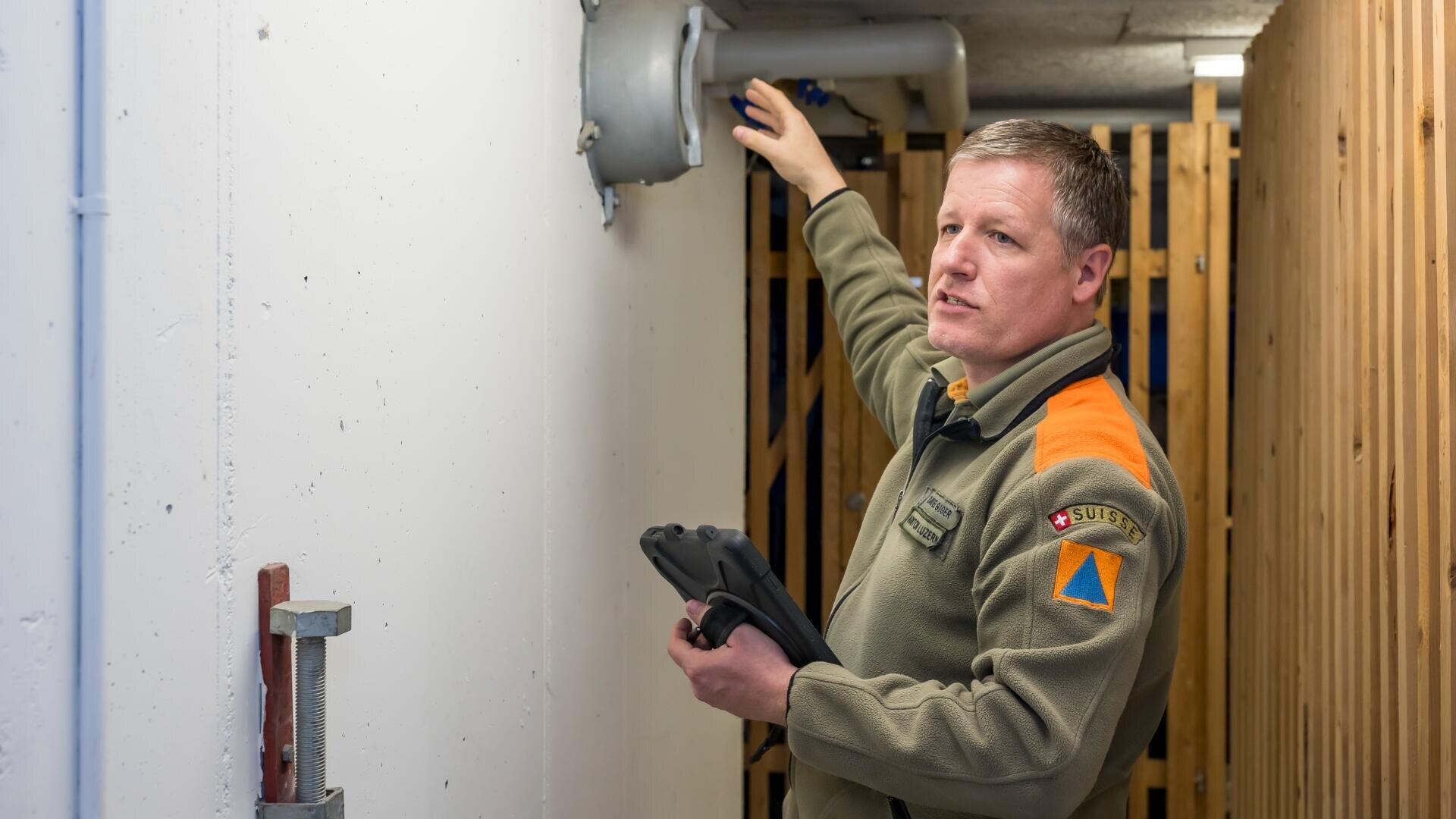 Ein Mann in grüner Uniform mit orangefarbenen Schulterabzeichen inspiziert eine graue Lüftungsanlage, die an einer weißen Wand montiert ist. Er hält ein Tablet in der einen Hand und deutet mit der anderen auf die Lüftungsöffnung.