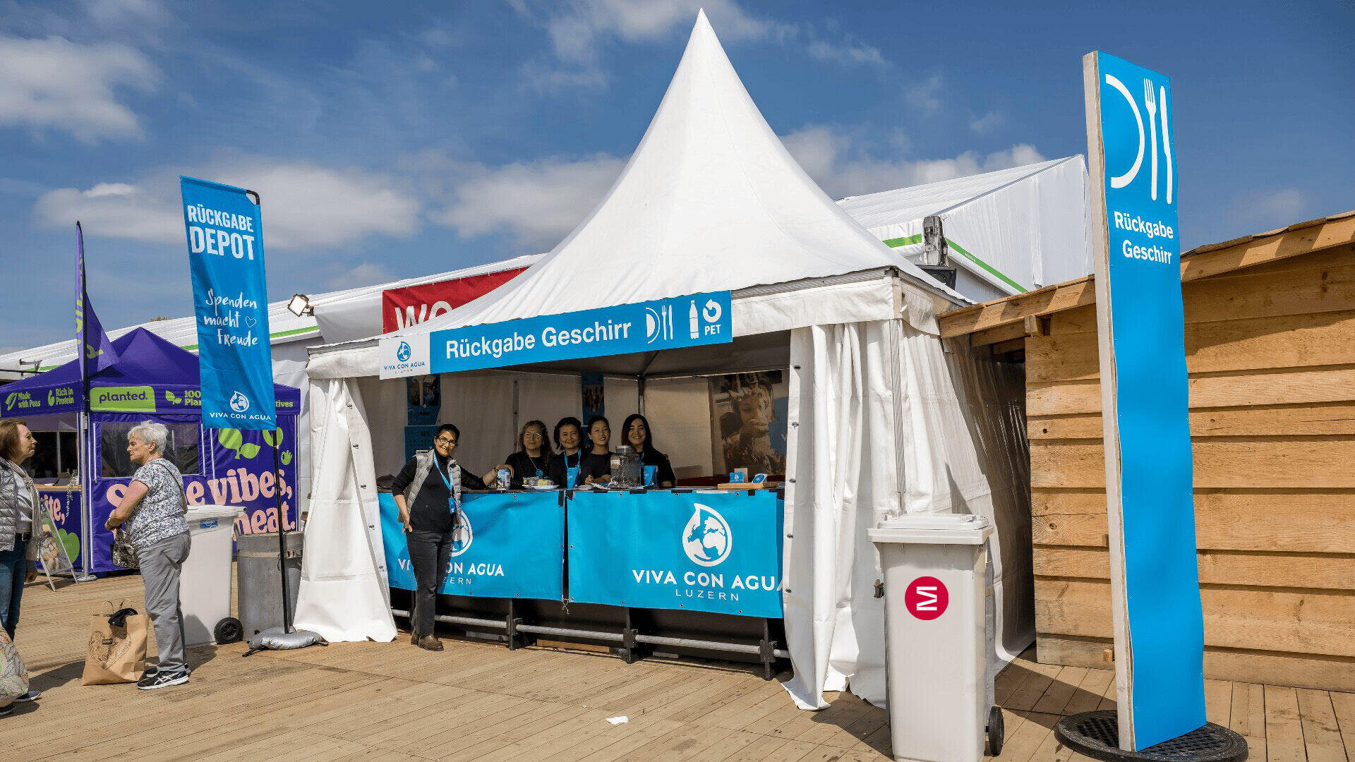 Ein weißes Zelt mit einem Schild "Rückgabe Geschirr" und dem Schriftzug "Viva con Agua" dient bei einer Veranstaltung im Freien als Station für die Rückgabe von Geschirr. Mehrere Personen stehen hinter dem Tresen, und ein paar Fußgänger laufen unter blauem Himmel in der Nähe.