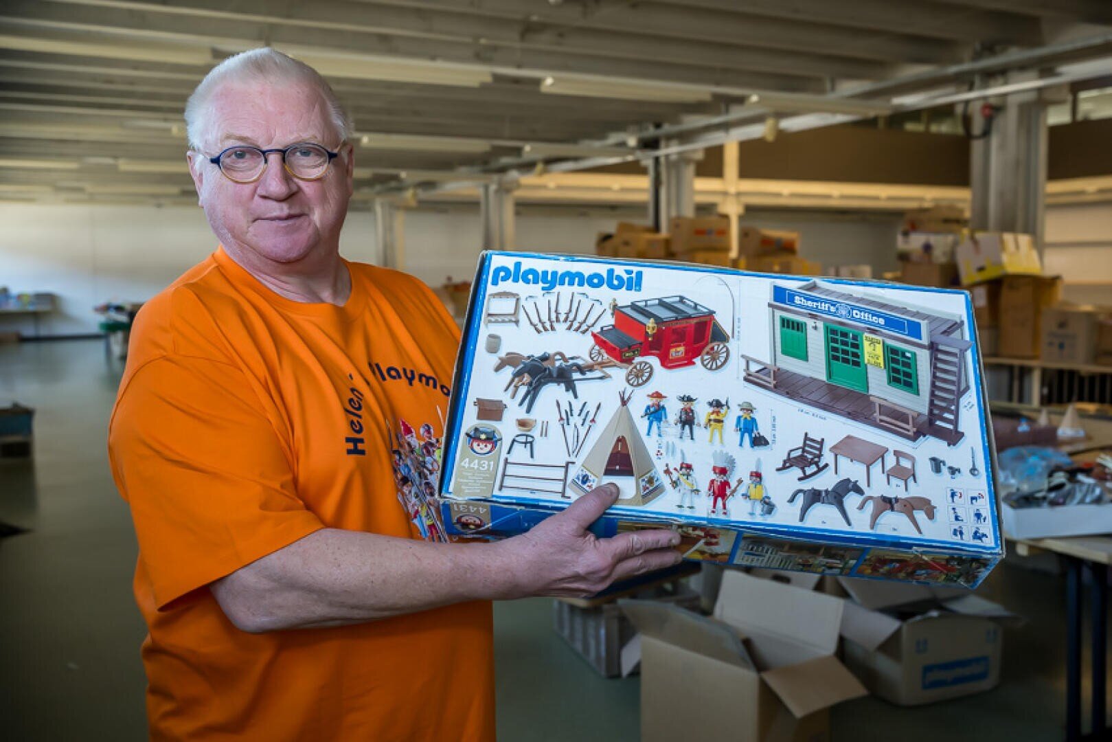 Ein älterer Mann mit Brille und orangefarbenem Hemd hält eine Playmobil-Spielzeugkiste mit einer Westernkutsche und Figuren. Er steht in einem großen Innenraum mit Kisten und Regalen im Hintergrund.