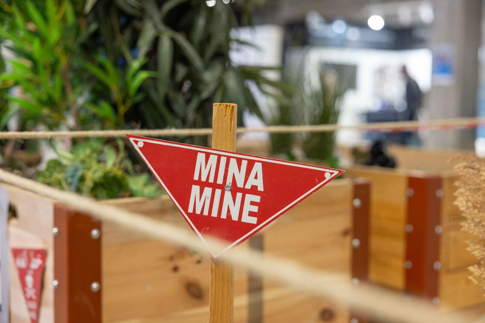 Ein rotes, dreieckiges Schild mit weißer Schrift, auf dem MINA MINE steht, ist auf einem Holzstab angebracht, neben abgesperrtem Grün und hölzernen Pflanzkübeln im Innenbereich.