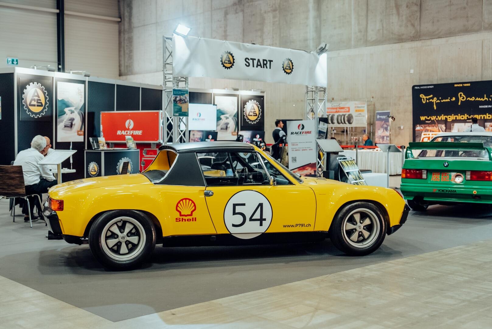 Ein gelber klassischer Rennwagen mit der Nummer 54 und dem Shell-Logo ist in einem Ausstellungsraum ausgestellt, mit Bannern, Ständen und einem weiteren grünen Auto im Hintergrund.