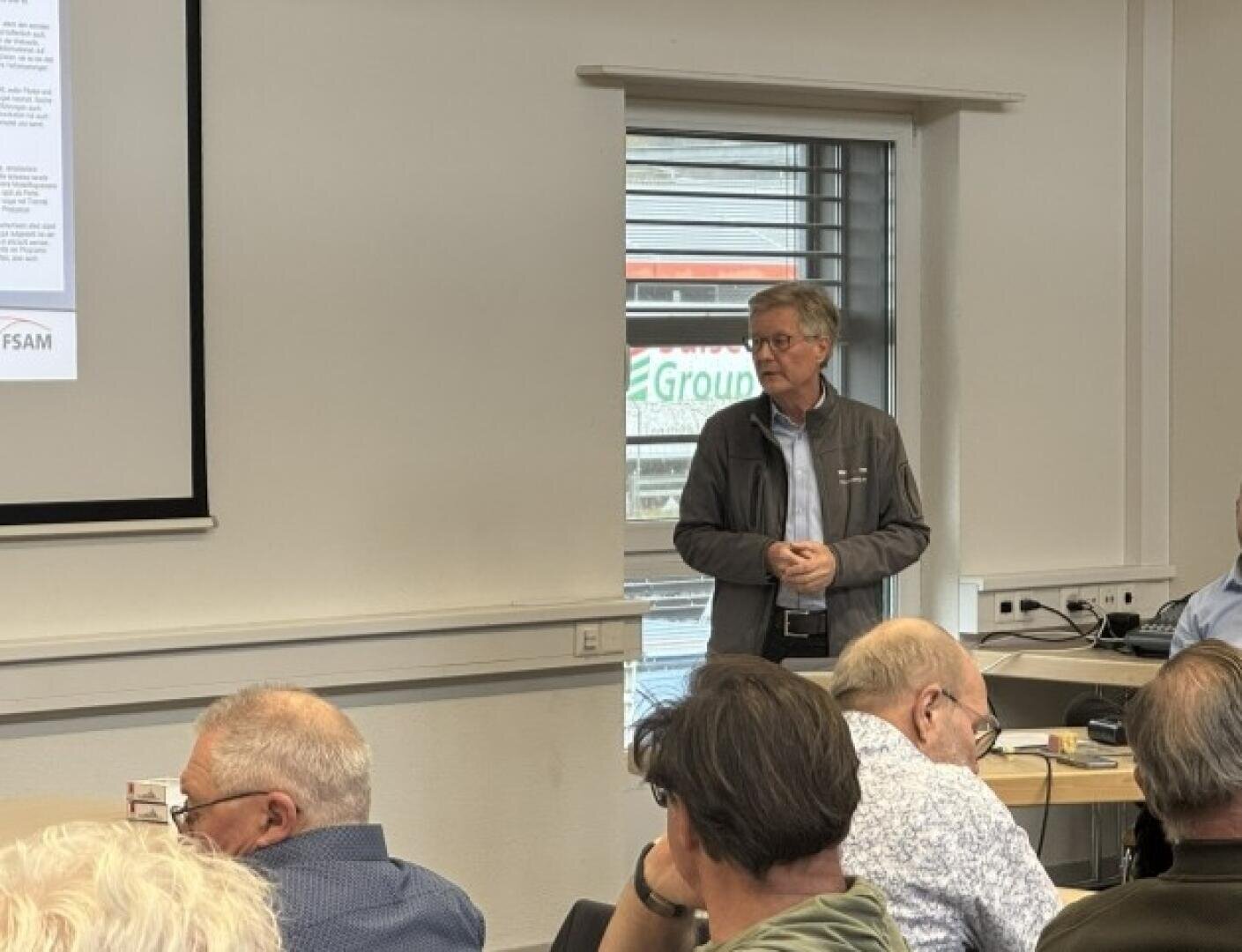 Ein Mann steht und spricht vor einer Gruppe von sitzenden Personen in einem Klassenzimmer oder Sitzungssaal, in der Nähe eines Fensters und einer projizierten Präsentationsfolie an der Wand.