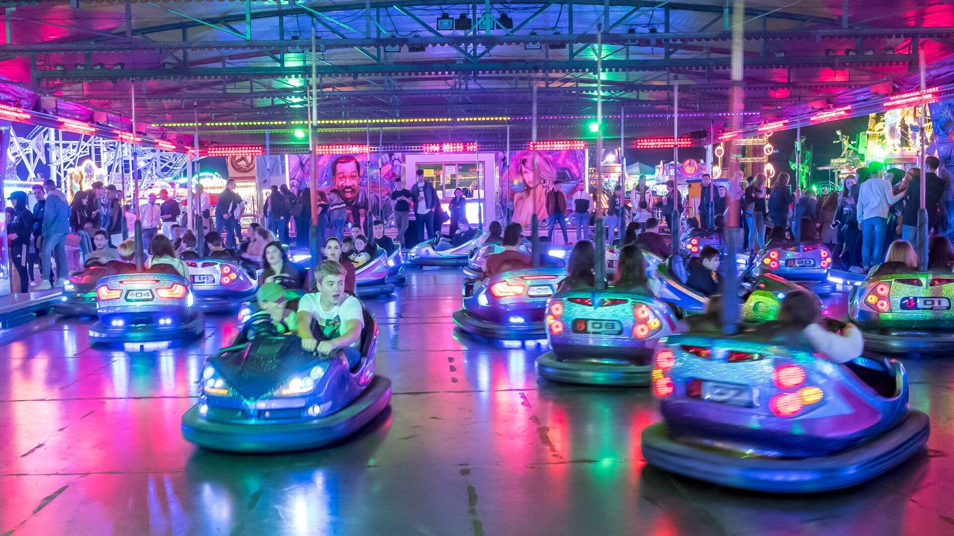 Menschen fahren bunte Autoscooter unter hellen Neonlichtern in einem belebten Indoor-Vergnügungspark, während eine Menschenmenge im Hintergrund zusieht. Die Szene ist lebendig, mit einer festlichen Atmosphäre und lebhaften Farben.