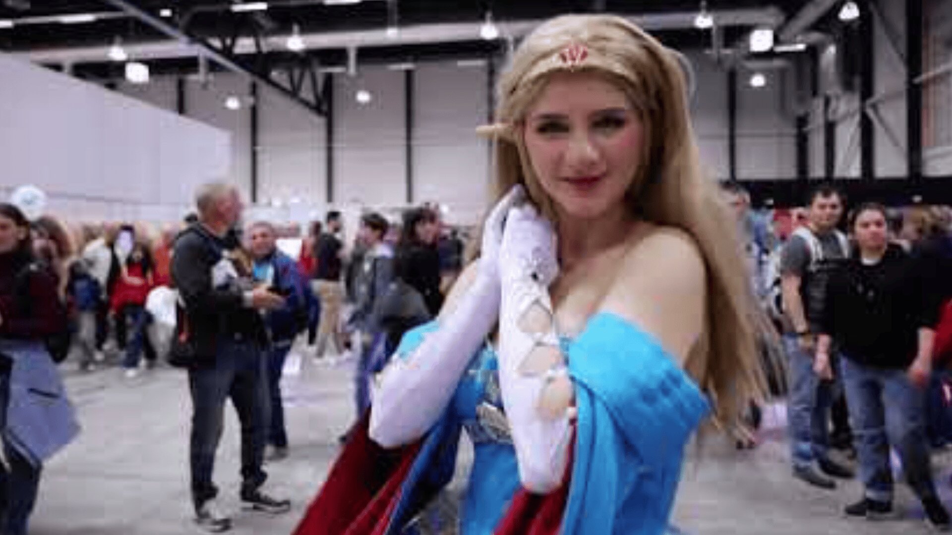 Eine Person in einem aufwendigen Fantasiekostüm posiert auf einer Convention; sie trägt Elfenohren, ein blau-rotes Kleid, weiße Handschuhe und eine Tiara, im Hintergrund sind eine Menschenmenge und Stände zu sehen.