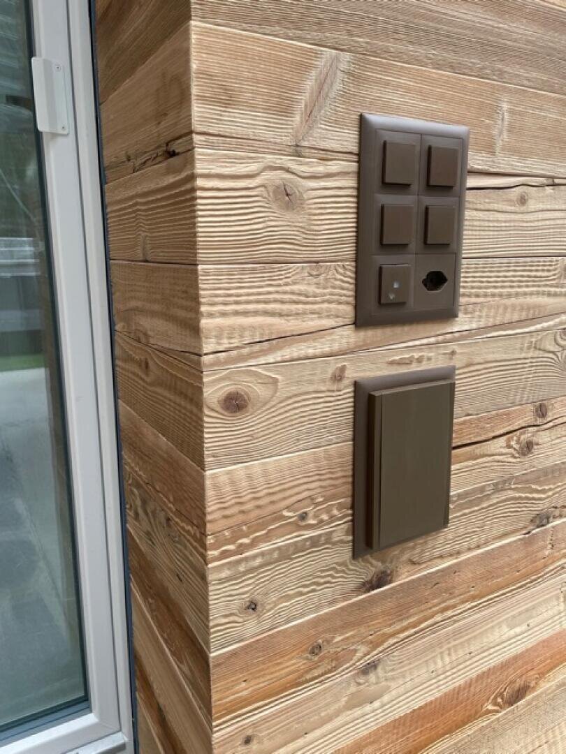 Braune Abdeckungen für Elektro- und Datensteckdosen sind an einer Wand mit horizontaler Holzverkleidung neben einer Glastür angebracht. Das Holz hat eine natürliche, strukturierte Maserung.