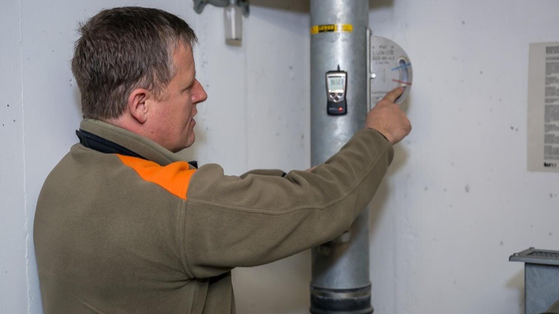 Eine Person in einer grün-orangefarbenen Jacke stellt ein Zifferblatt an einem großen vertikalen Rohr in einem Innenraum ein. An dem Rohr ist ein digitales Messgerät befestigt, und an der Wand daneben hängt ein Blatt Papier.