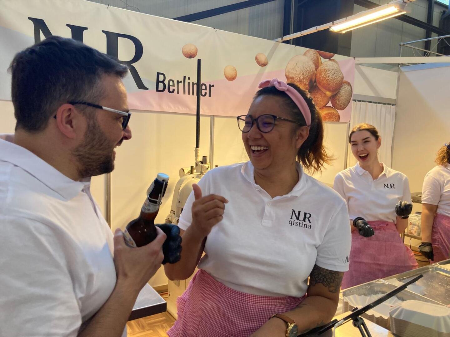 Vier Personen in weißen Hemden und rosa Schürzen stehen an einem Essensstand mit der Aufschrift NR Berliner. Zwei Personen im Vordergrund lächeln und unterhalten sich, eine hält ein Getränk in der Hand. Im Hintergrund sind Gebäck und Backgeräte zu sehen.