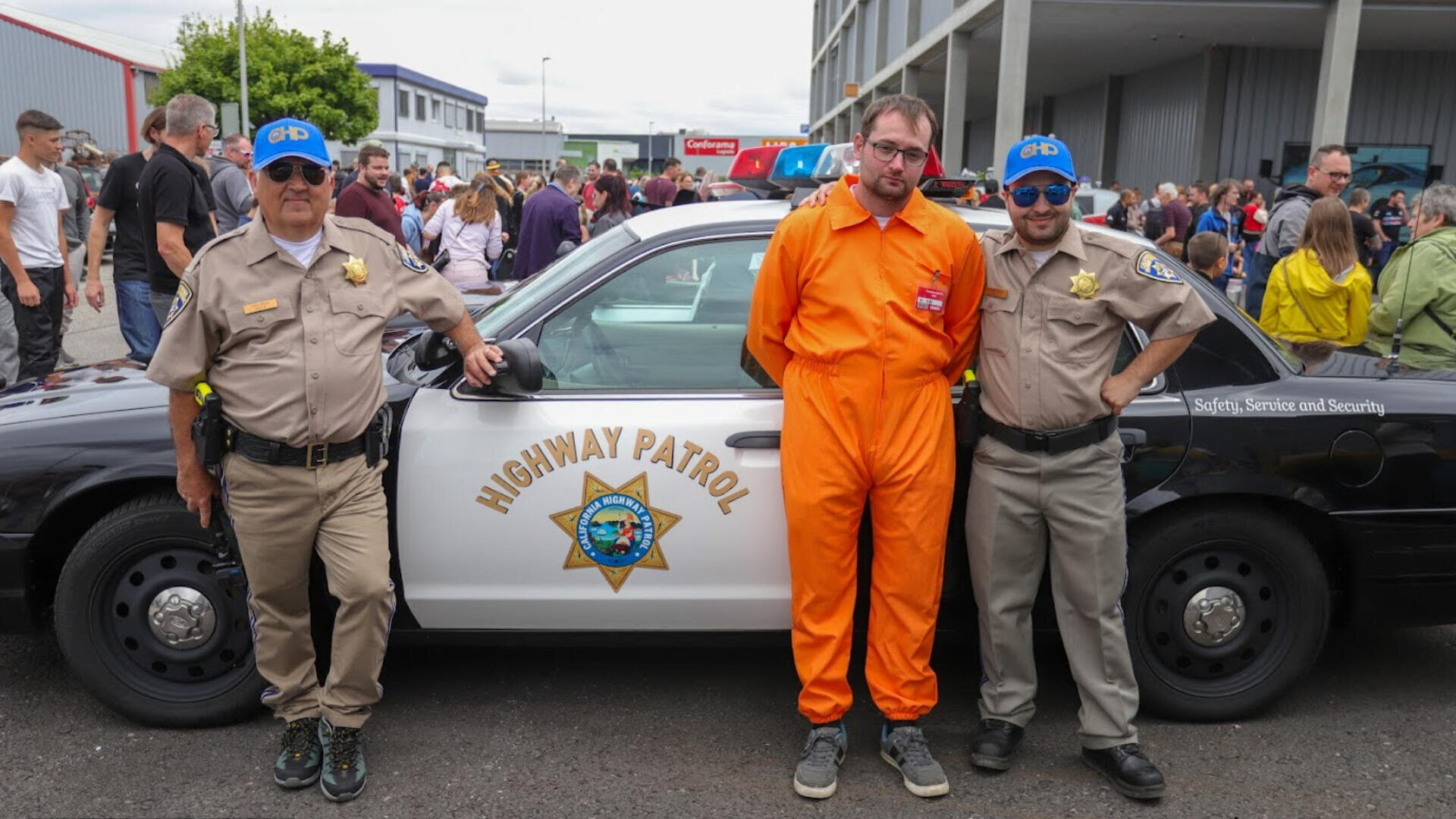 Drei Männer posieren vor einem Auto der Highway Patrol. Zwei tragen hellbraune Polizeiuniformen und blaue Baseballmützen, während der Mann in der Mitte einen orangefarbenen Overall trägt. Im Hintergrund ist eine Menschenmenge zu sehen.