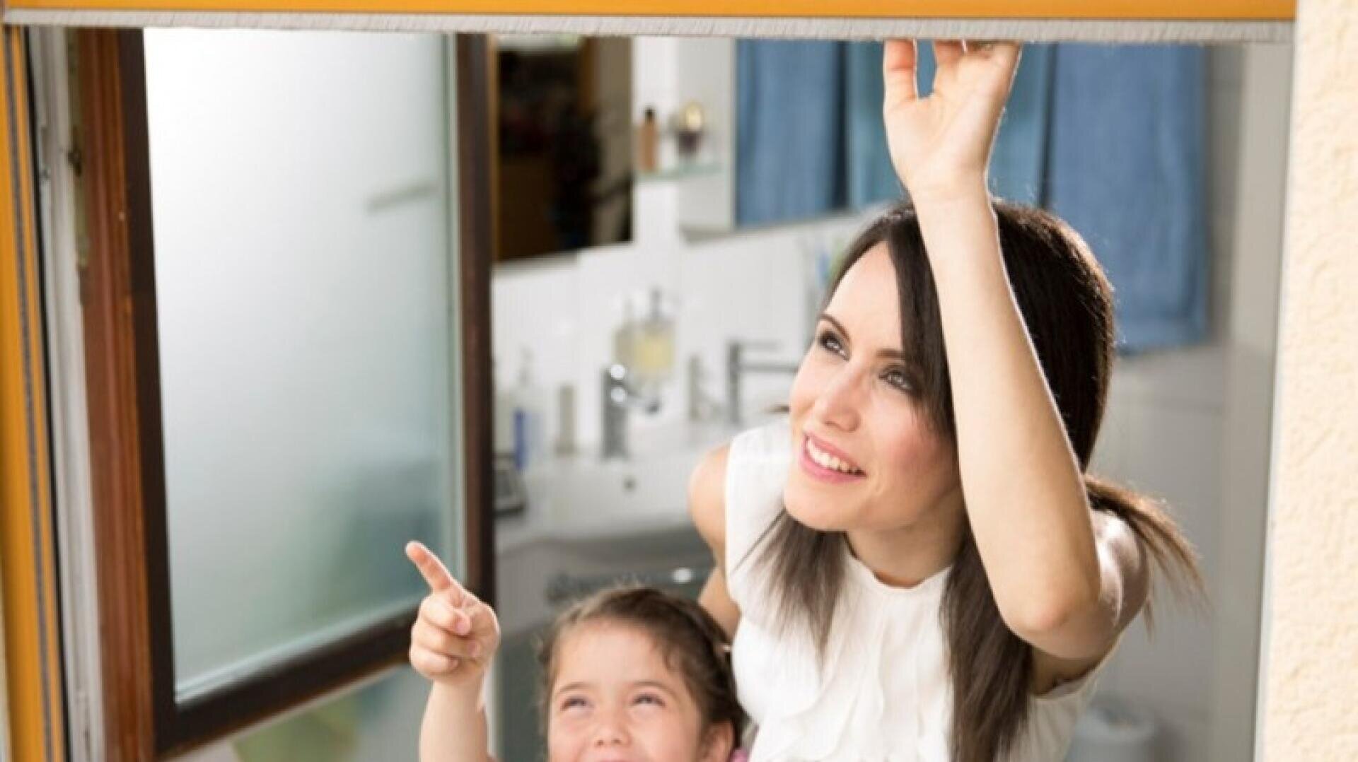 Eine Frau und ein junges Mädchen lächeln, während sie eine Fensterscheibe öffnen. Die Frau hebt die Scheibe hoch, und das Mädchen zeigt nach draußen, wobei beide fröhlich aussehen. Die Szene findet in einem Innenraum in der Nähe eines Badezimmers statt.