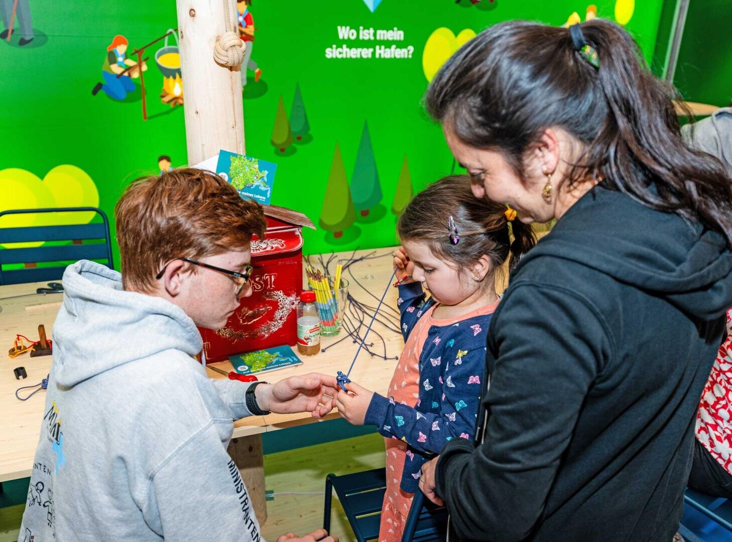 Ein Mann und eine Frau helfen einem jungen Mädchen bei einer Bastelarbeit an einem Tisch. Der Hintergrund ist hellgrün mit Illustrationen und deutschem Text. Das Mädchen ist auf das Projekt konzentriert, während die Erwachsenen ihr helfen.