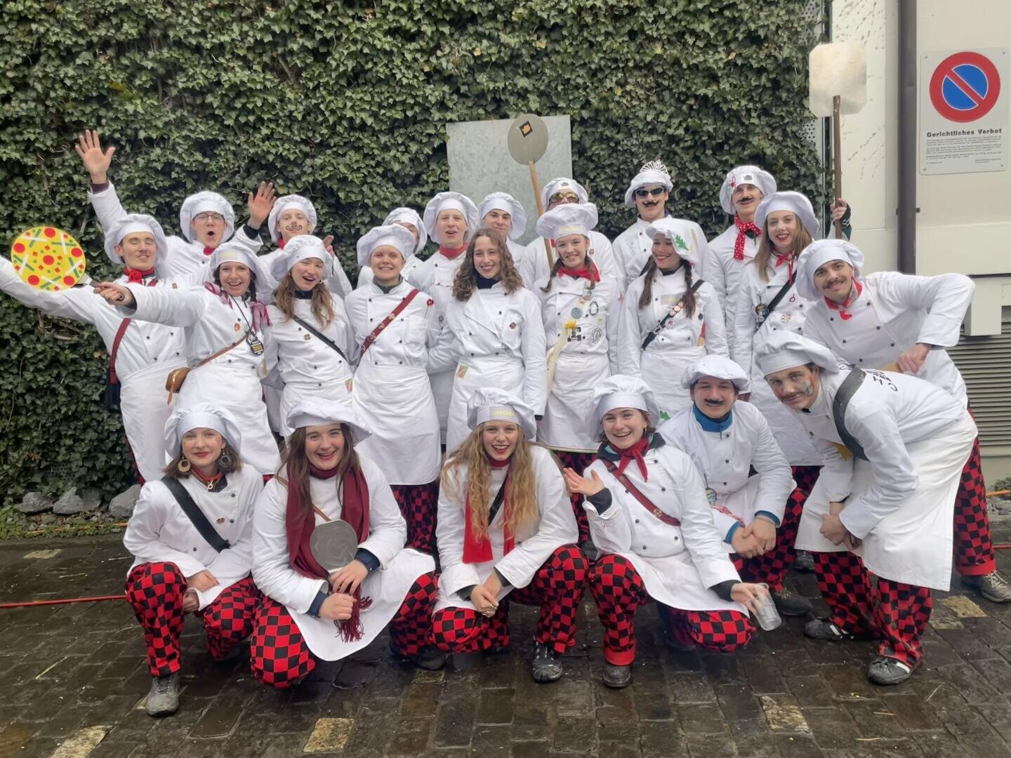 Eine Gruppe lächelnder, als Köche gekleideter Menschen mit weißen Mützen und Jacken mit rot karierten Hosen posiert gemeinsam im Freien auf einer Kopfsteinpflasterstraße, mit einer efeubewachsenen Mauer im Hintergrund. Einige halten Küchenutensilien in der Hand.