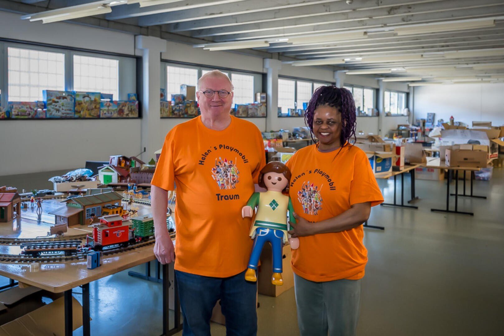 Zwei Personen in orangefarbenen Hemden mit Text stehen in einem Raum voller Spielzeugeisenbahnen und Playmobil-Sets. Eine Person hält eine große Playmobil-Figur in der Hand. Der Raum ist durch große Fenster hell erleuchtet.