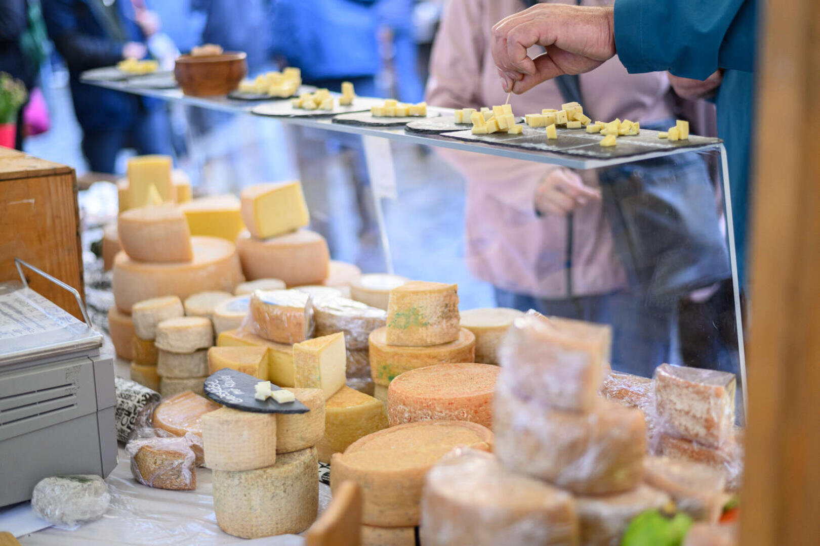 Auf einem Marktstand sind verschiedene runde Käsesorten ausgestellt, auf denen Tabletts mit Käseproben stehen. Die Leute strecken ihre Hände aus, um Käseproben zu nehmen, und im Hintergrund herrscht reges Treiben mit Einkäufern.