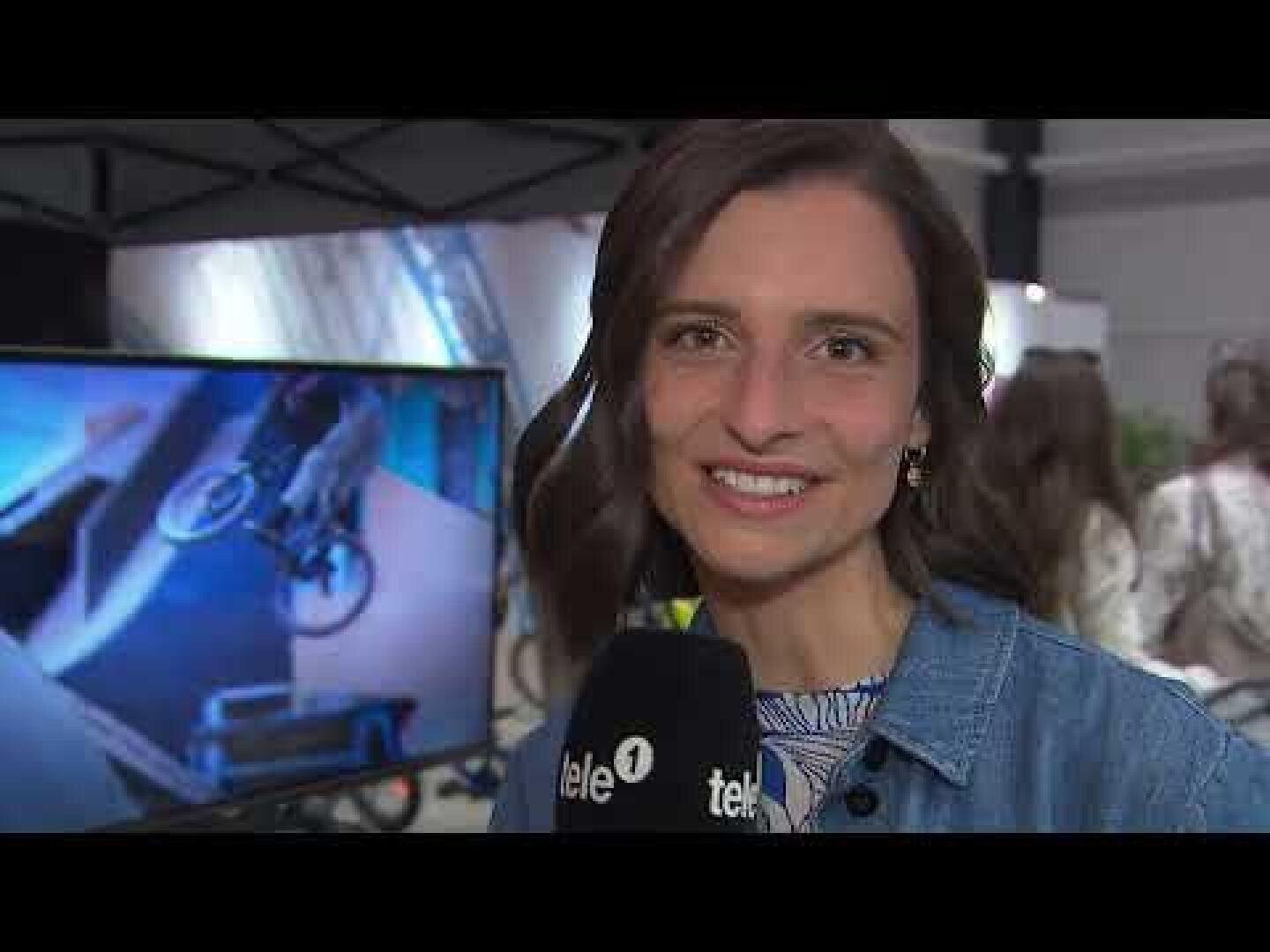 Eine Frau lächelt in die Kamera und hält ein Mikrofon mit der Aufschrift tele. Hinter ihr zeigt ein Fernsehbildschirm eine Person, die einen BMX-Trick vorführt, und im Hintergrund sind mehrere Personen versammelt.