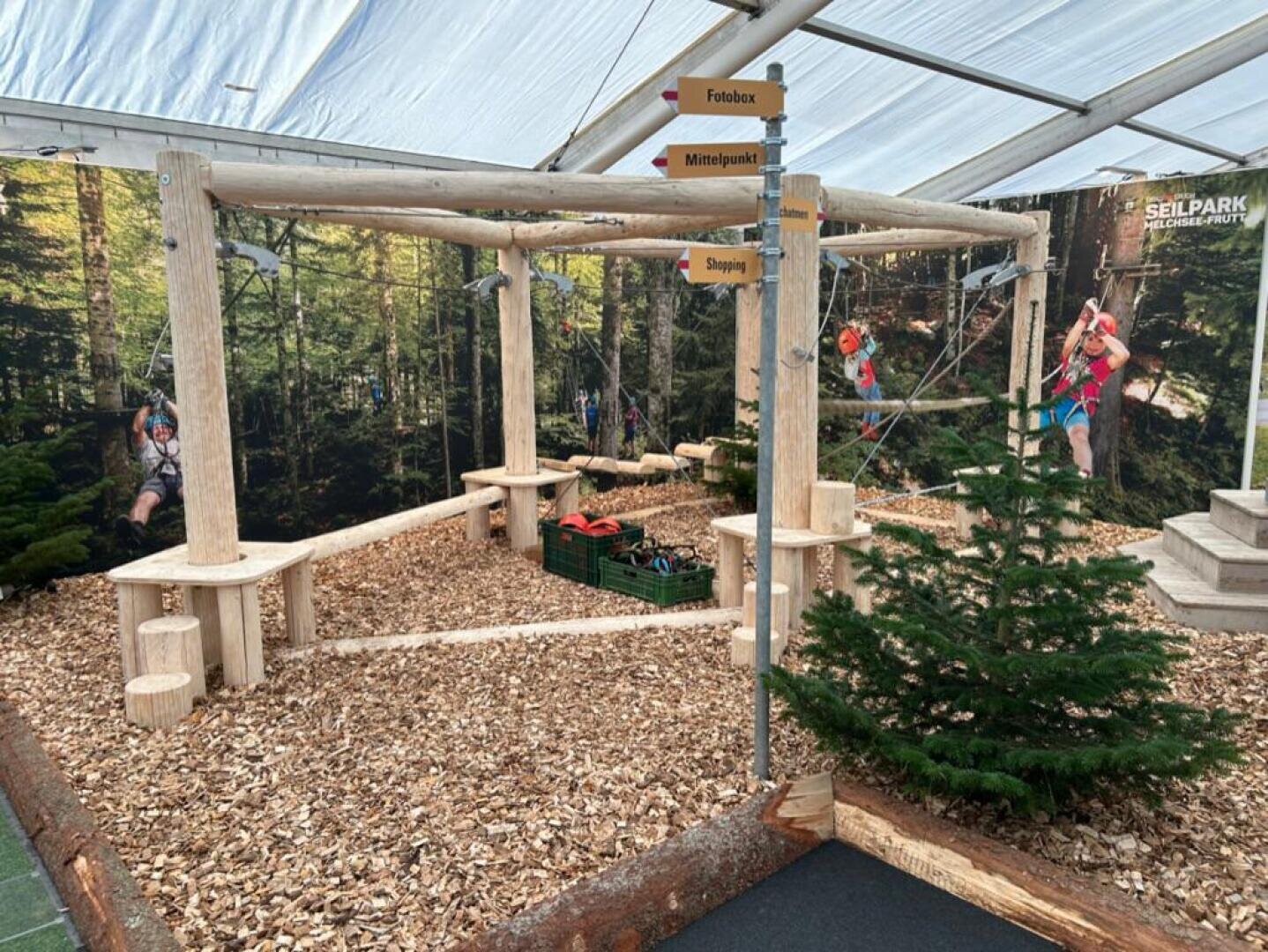 Ein kleiner Indoor-Hindernisparcours aus Holz mit Bänken, Mulchböden und Kletterausrüstung. Wegweiser weisen auf die verschiedenen Aktivitäten hin. Eine Waldszene und Kletterbilder schmücken die Wände im Hintergrund.