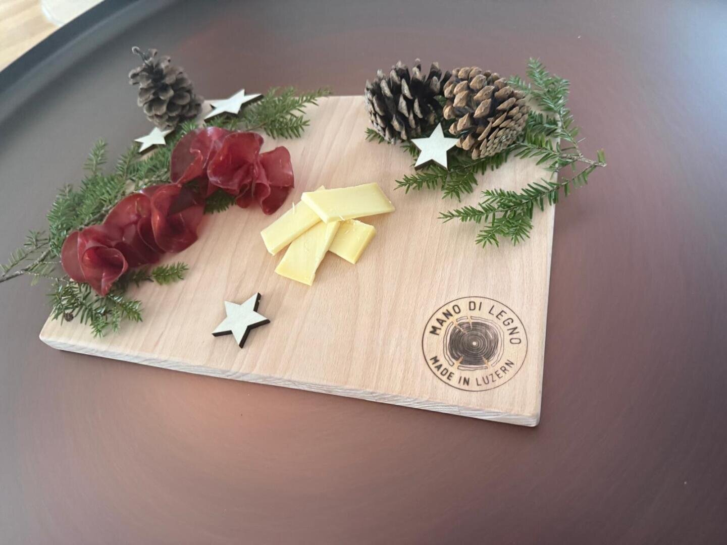 Ein Holzbrett mit Käsescheiben, gerolltem Wurstfleisch, Pinienzapfen, Tannenzweigen und Papiersternen, dekorativ angeordnet. Das Brett ist mit der Aufschrift "MANO DI LEGNO MADE IN LUCERNE" versehen.