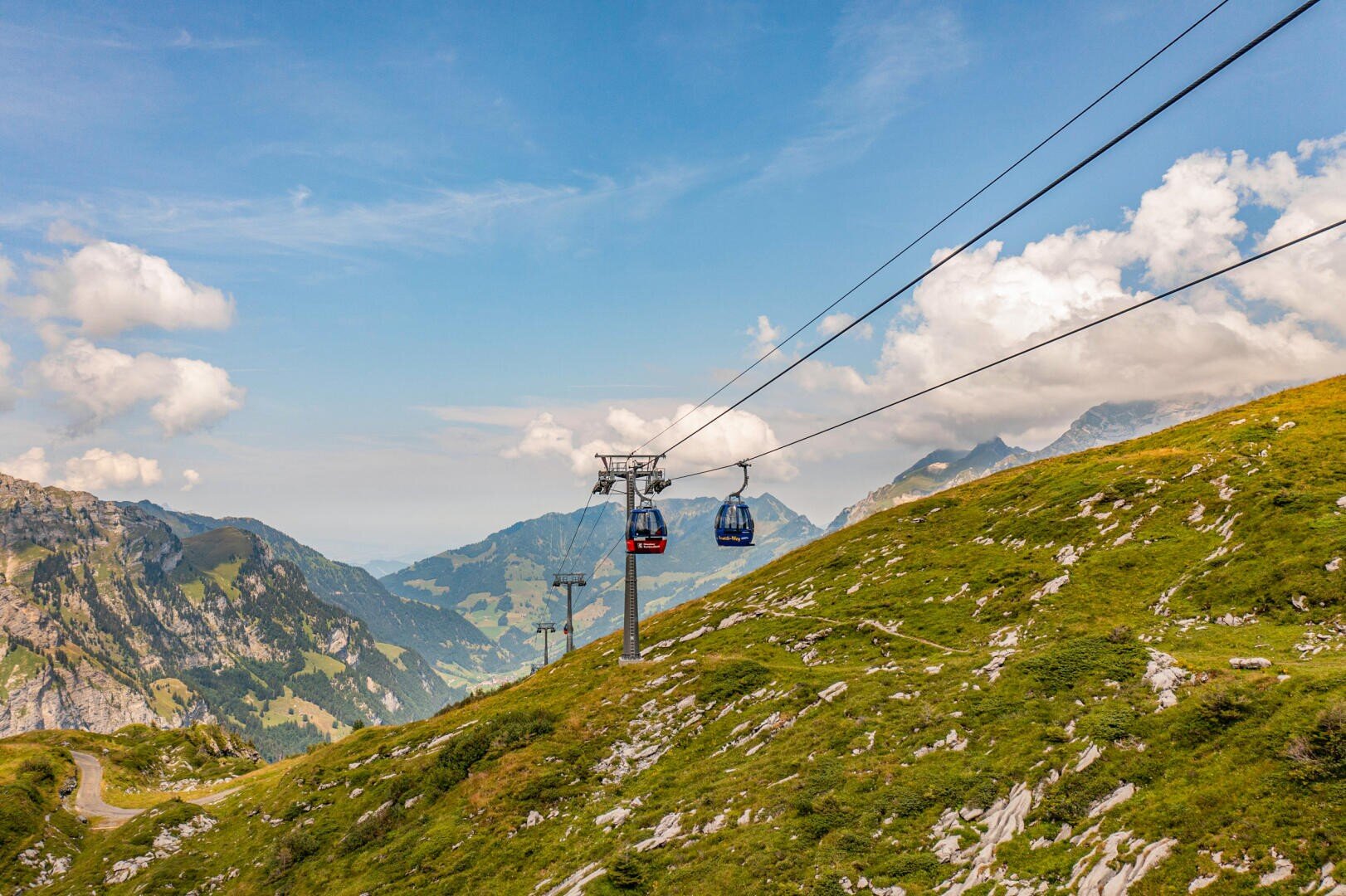 Seilbahnen fahren einen grünen, felsigen Berghang unter einem blauen Himmel mit vereinzelten Wolken hinauf. Im Hintergrund sind majestätische Berge und ein Tal zu sehen.