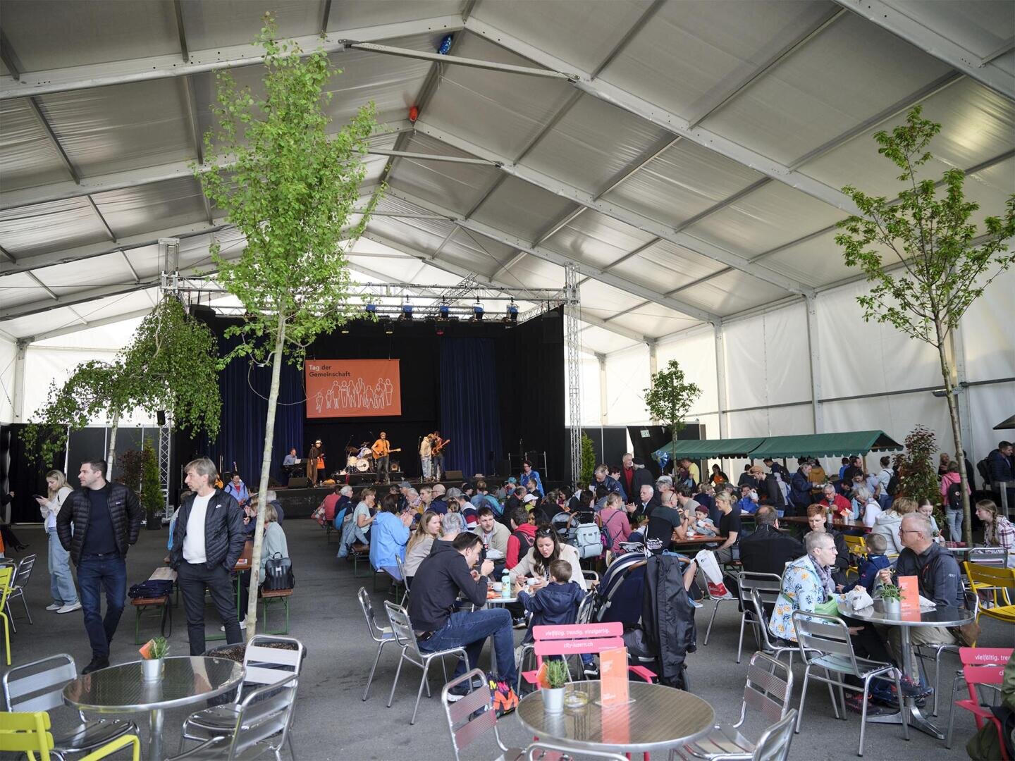 Menschen sitzen an Tischen, essen und unterhalten sich unter einem großen weißen Zelt mit Bäumen im Inneren. Auf einer Bühne im vorderen Bereich spielt eine Band. Es herrscht eine lebhafte und gesellige Atmosphäre, in der sowohl Erwachsene als auch Kinder anwesend sind.