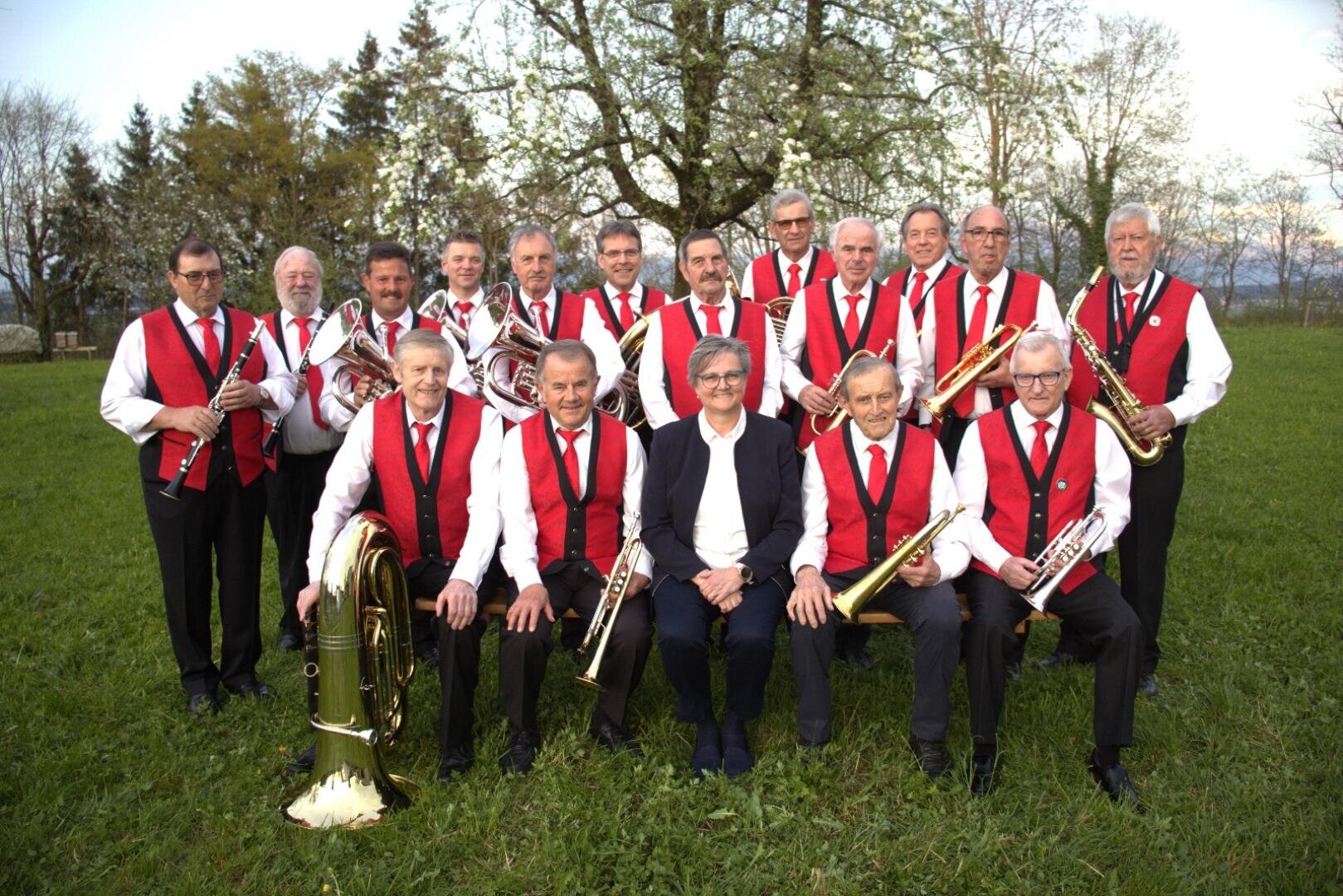 Eine Blaskapelle von 17 Männern, die meisten in weißen Hemden, schwarzen Hosen und roten Westen, posiert im Freien auf einer Wiese vor Bäumen, hält verschiedene Blasinstrumente in der Hand und lächelt in die Kamera.